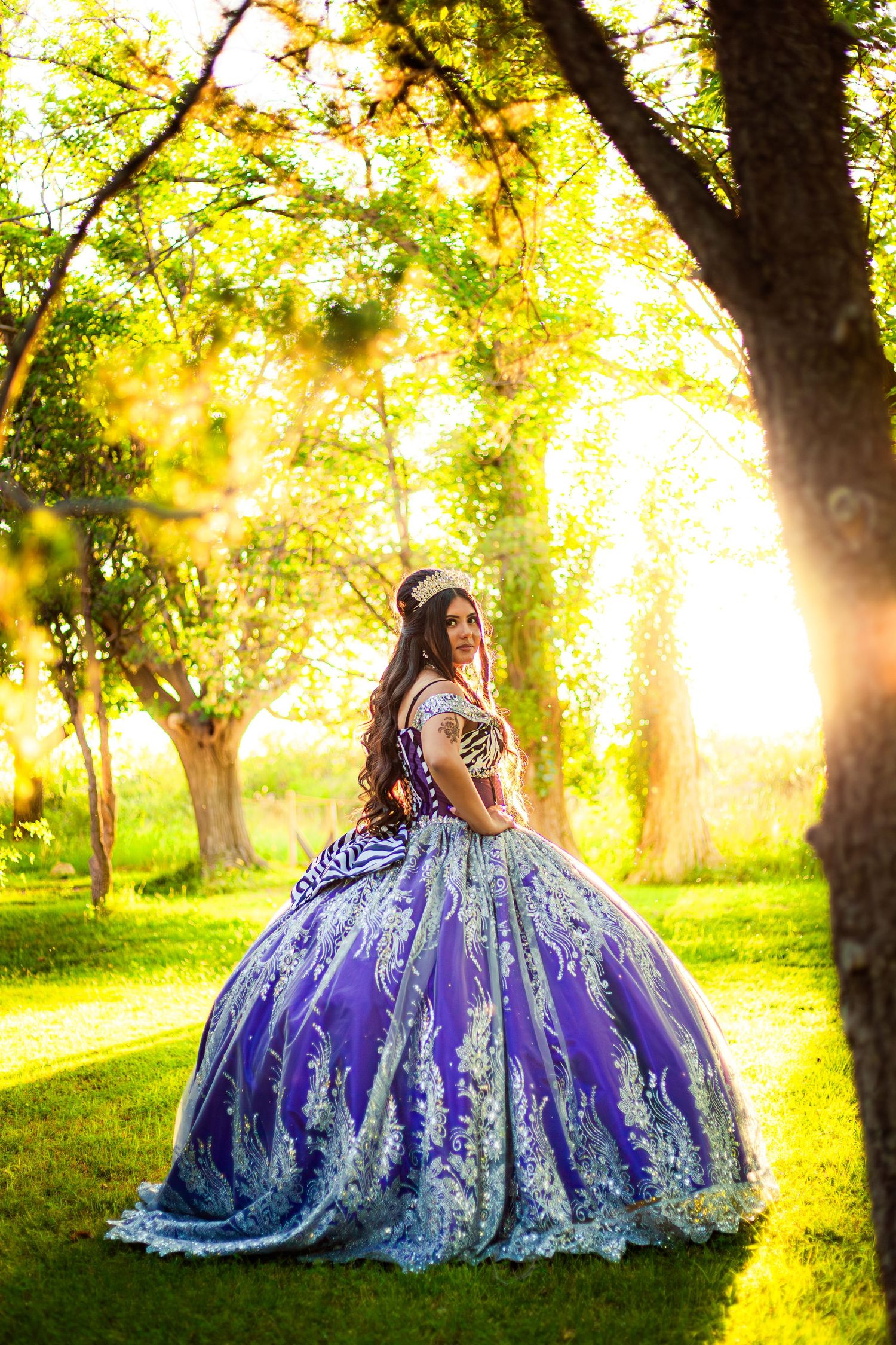 Young woman in purple ball gown with white lace posing in sunlit garden for quinceañera.