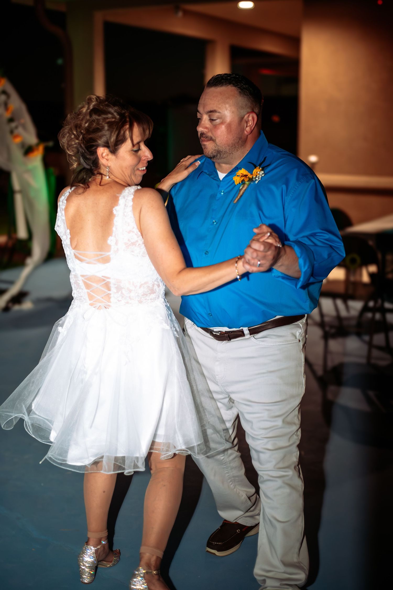 Bride and groom share first dance, bride in white dress, groom in blue shirt with sunflower boutonniere.