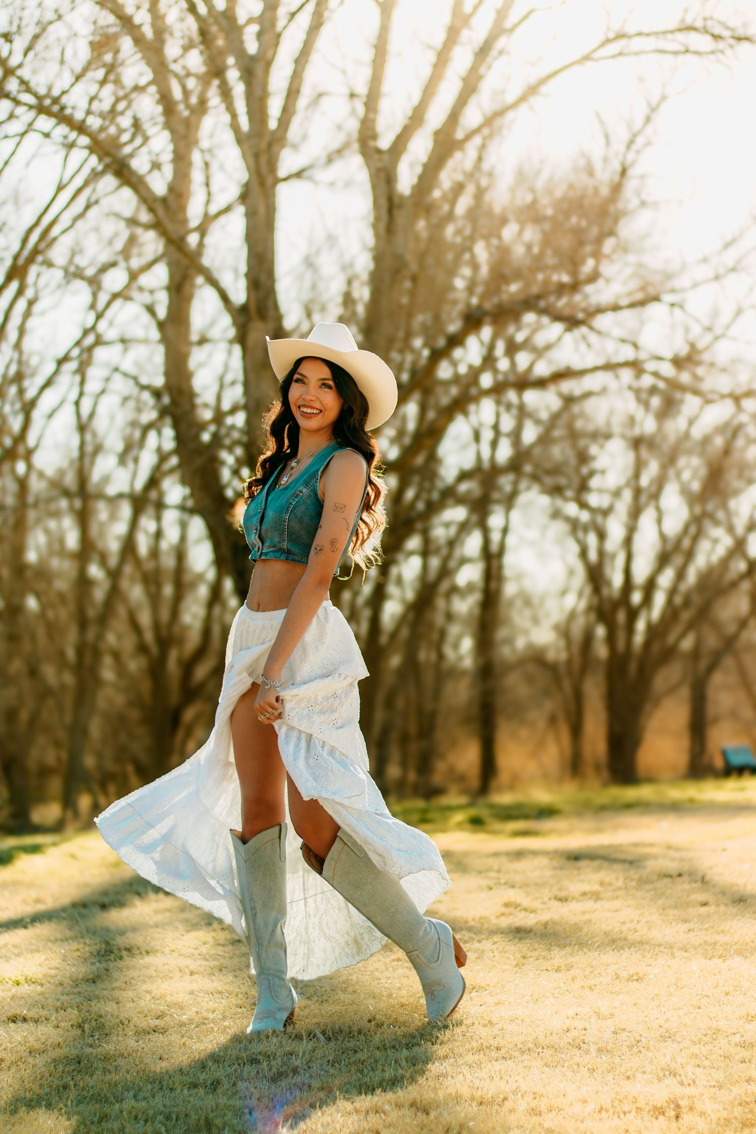 Smiling brunette in teal crop top, white skirt and cowboy boots twirls in golden sunlit field.