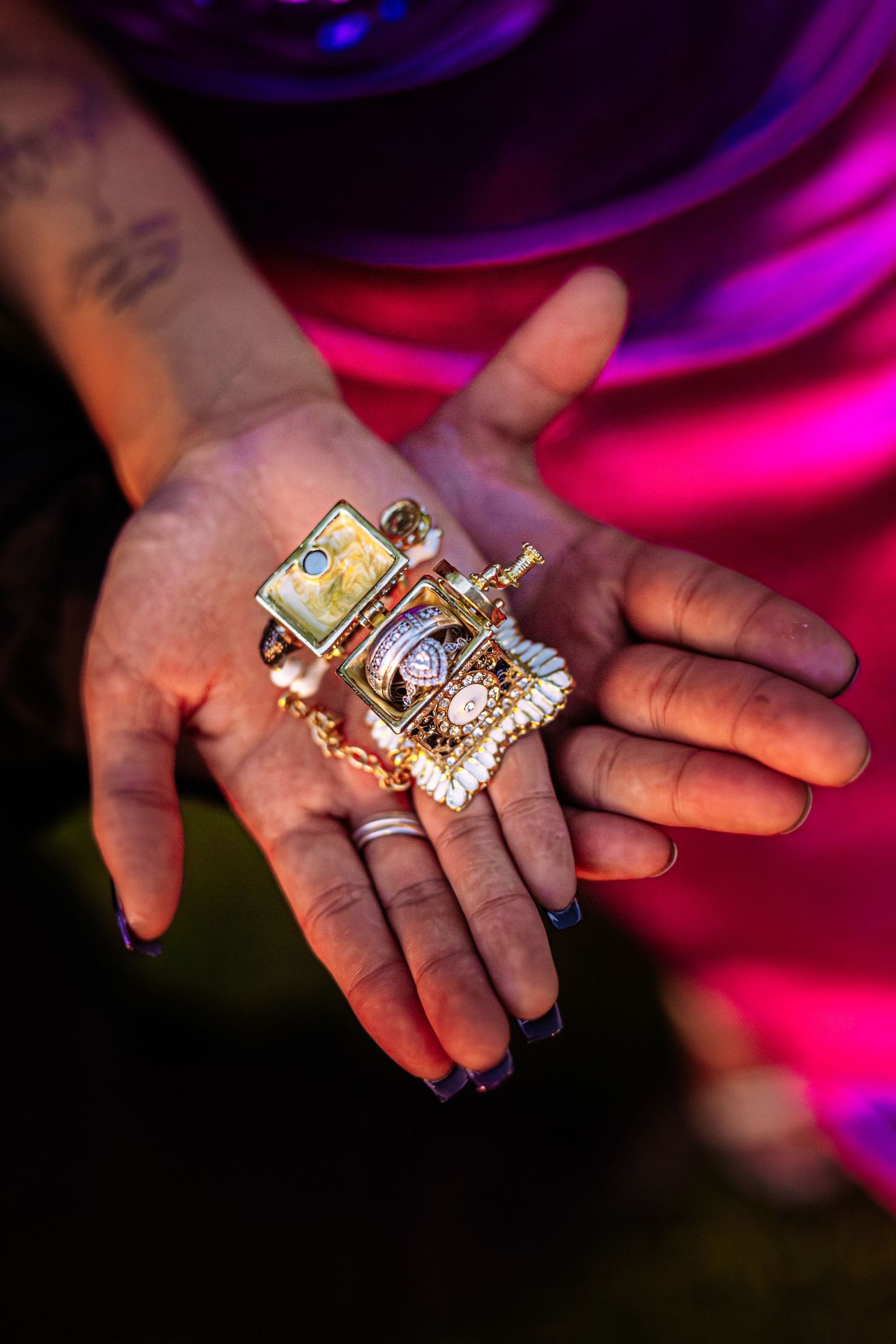 Tattooed hands holding ornate gold jewelry pieces under vibrant pink and purple festival lights.