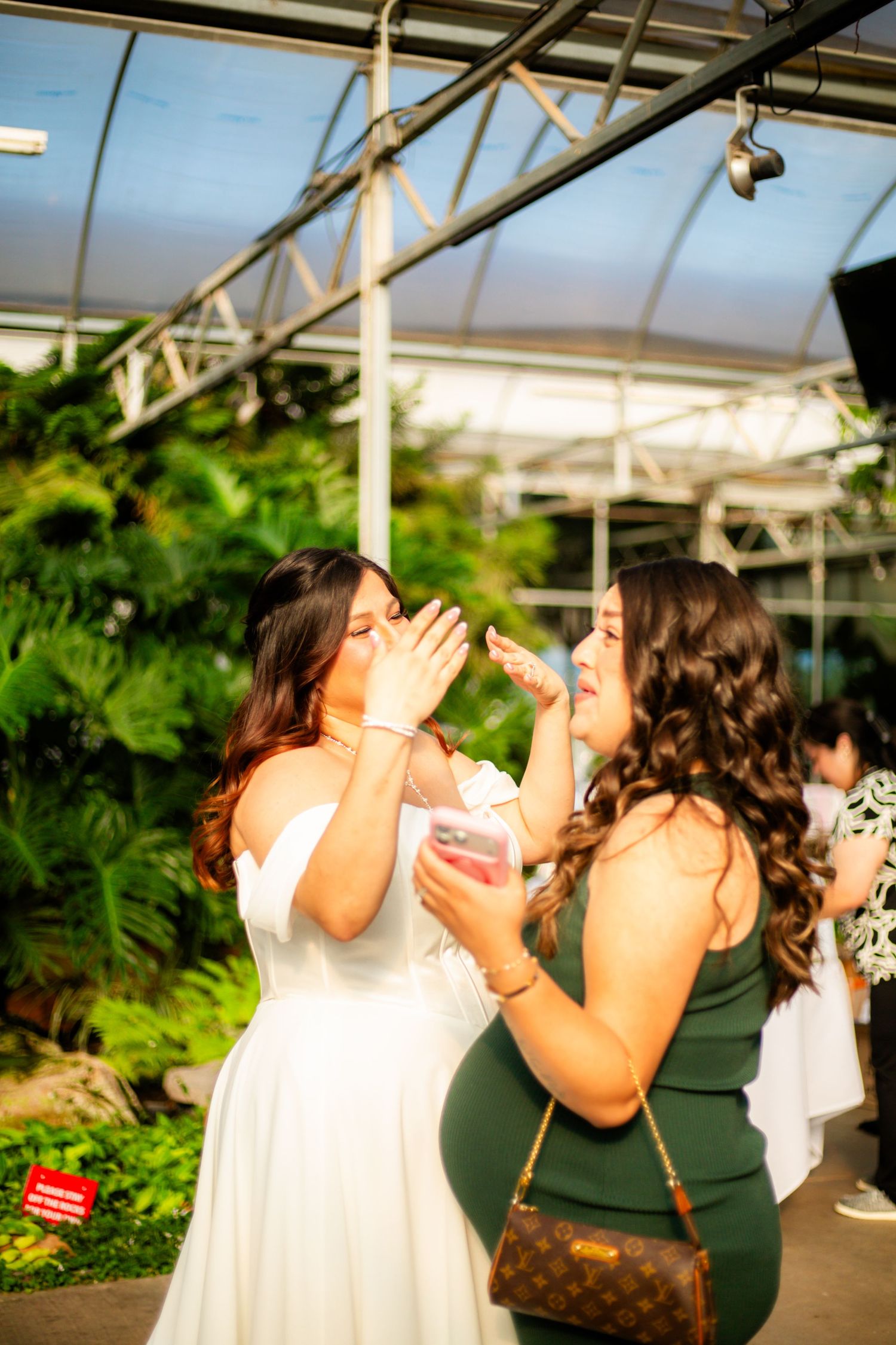 Two women in a greenhouse venue, one in white bridal dress and one in green, sharing a sweet moment with dessert.