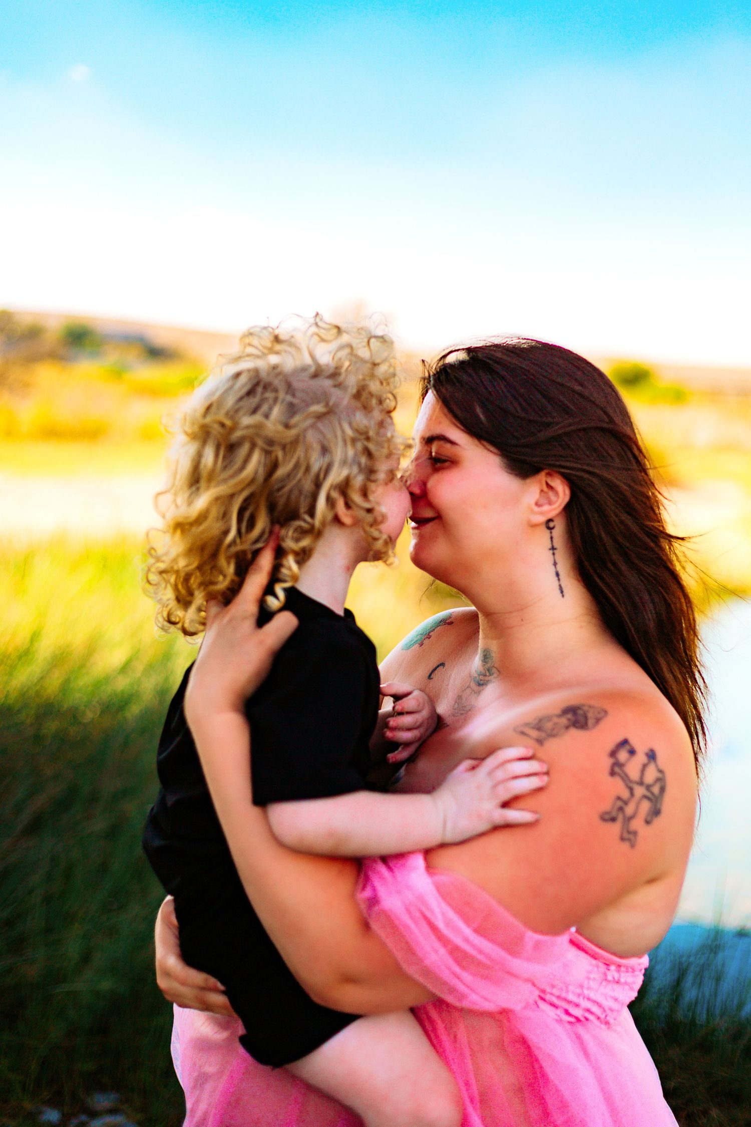 A tattooed mother in a pink dress holds her curly-haired toddler, sharing an Eskimo kiss outdoors.