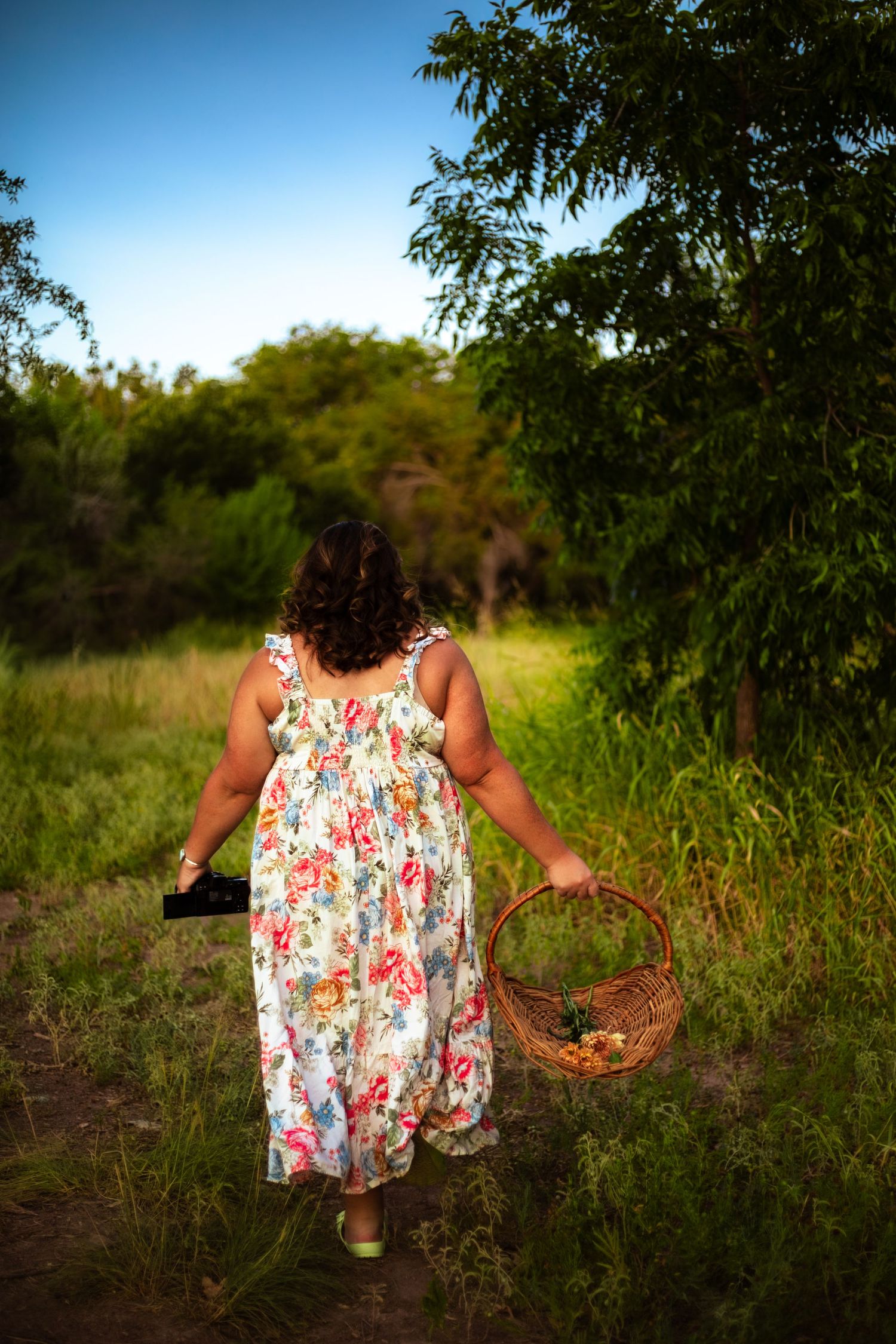 Woman in floral dress carrying wicker basket walking through lush green garden path on sunny day.