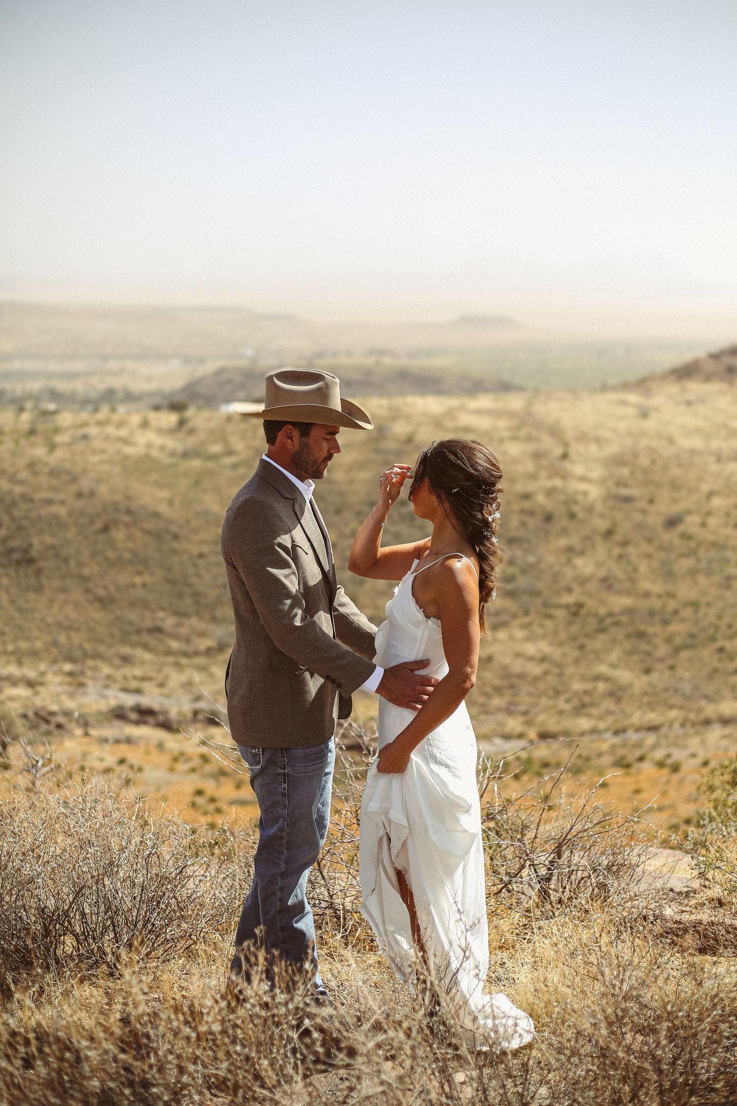 Couple in western attire sharing a romantic moment in a desert landscape, groom in cowboy hat and bride in white dress.