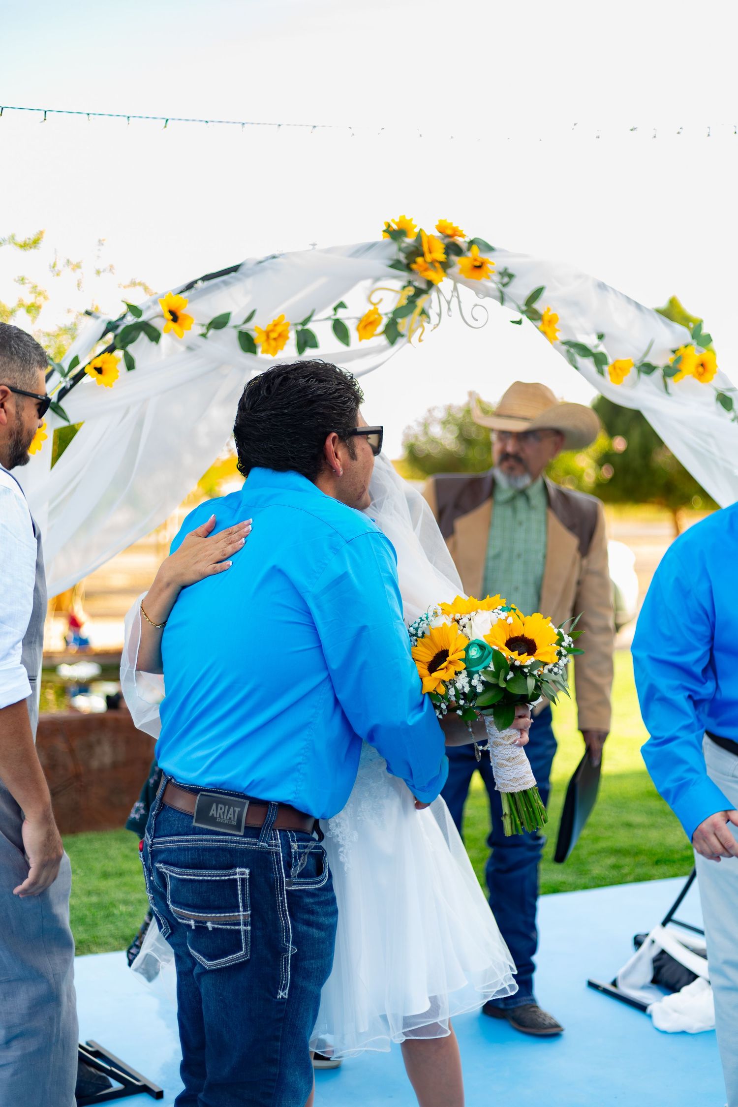 Groom in blue shirt embraces bride under sunflower arch at outdoor wedding ceremony.