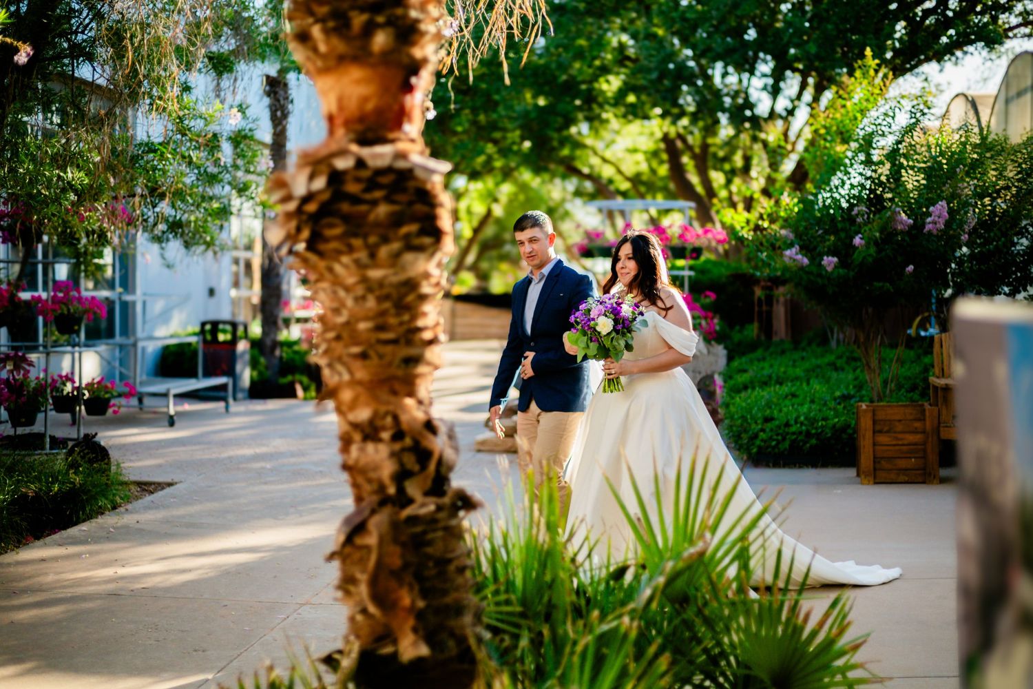 Bride and groom walking through a lush garden pathway on their wedding day, surrounded by colorful flowers and greenery.