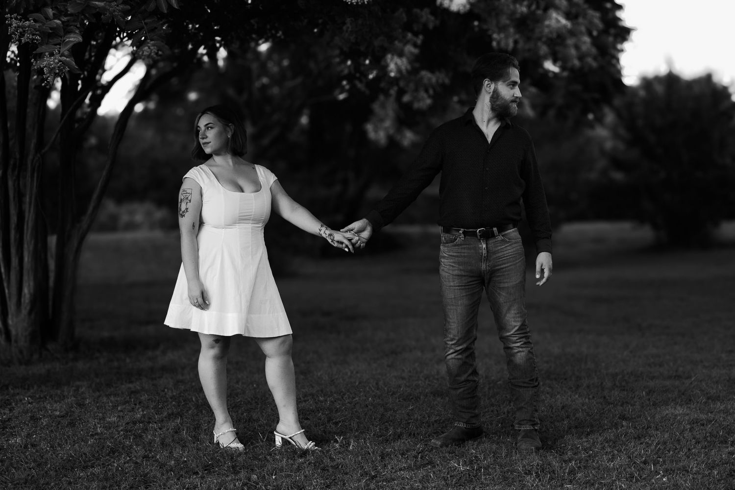 Black and white photo of couple holding hands in park at dusk under trees.