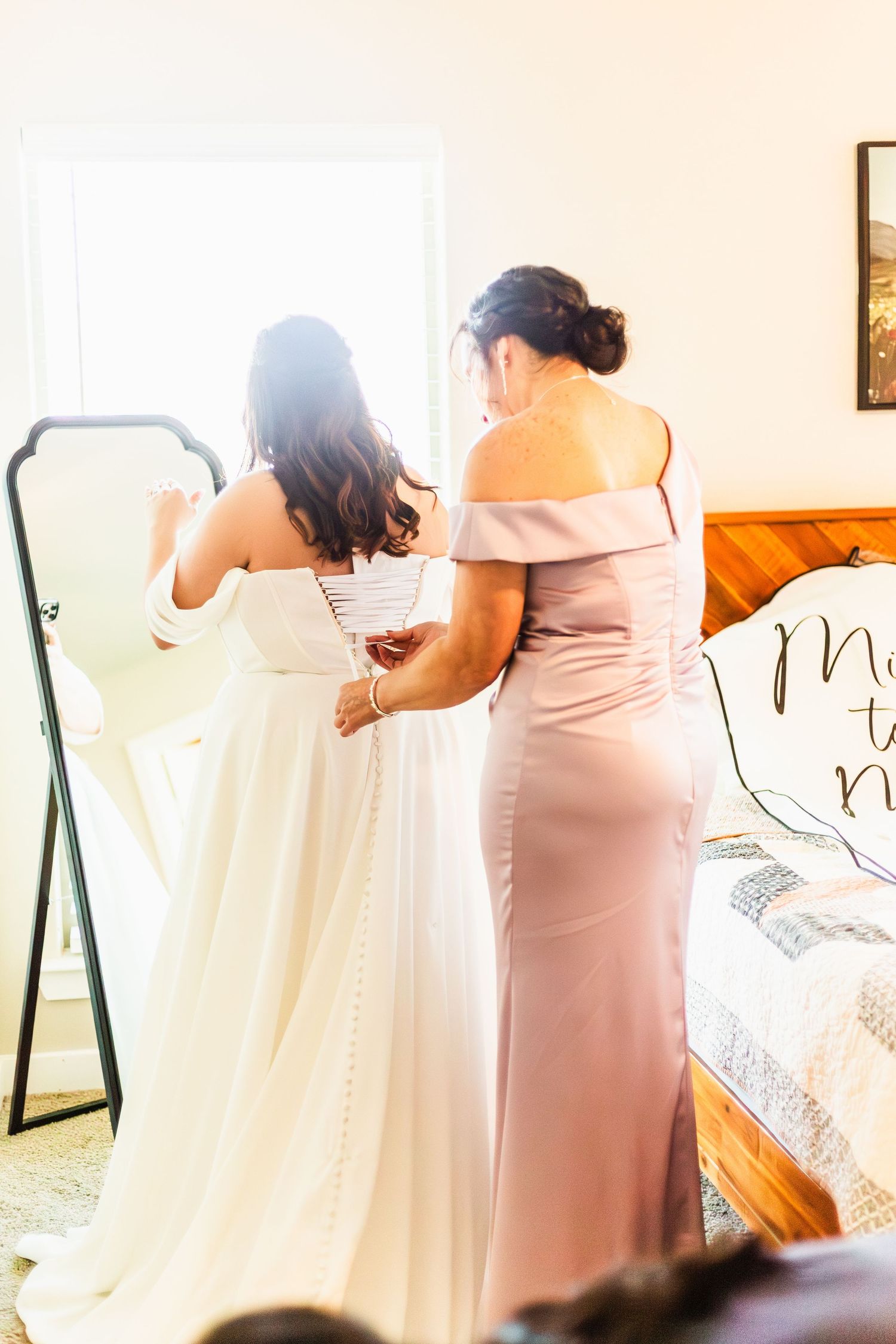Bridesmaid in blush gown helps bride button her white wedding dress in a sunlit bedroom beside a floor mirror.