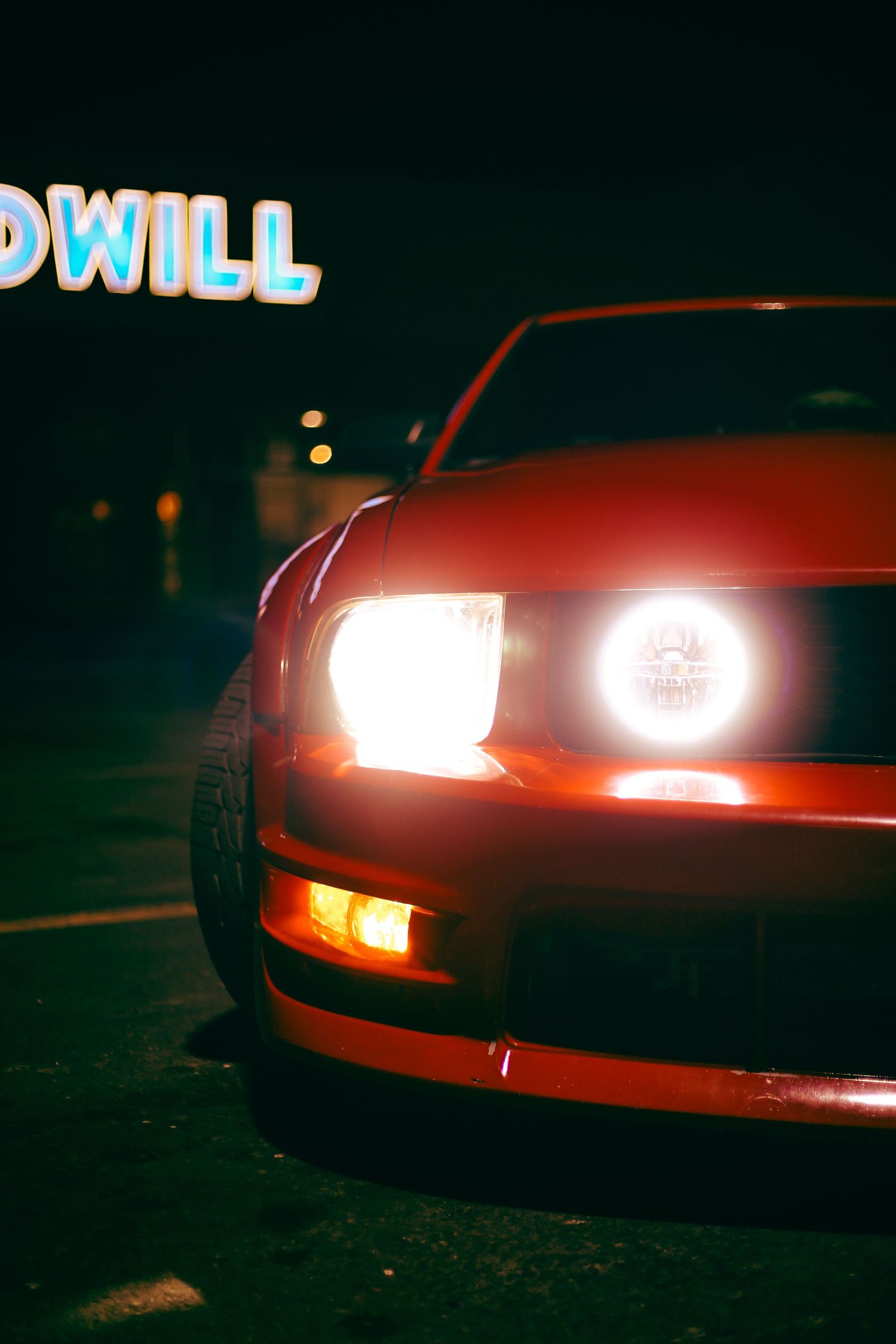 Red Ford Mustang with bright headlights illuminated at night near a Goodwill store sign.