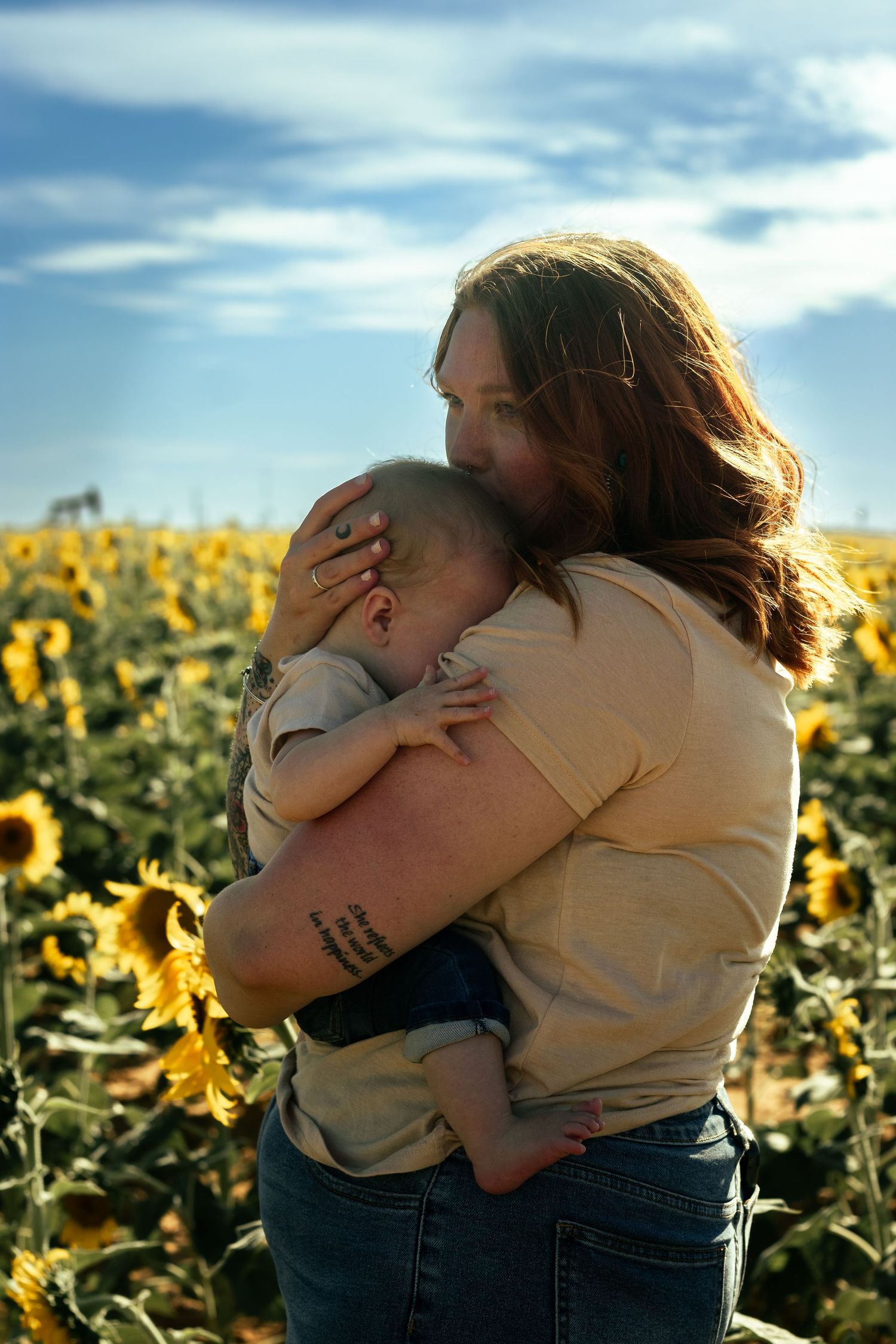 Mother embracing baby in golden sunflower field under dramatic cloudy sky.