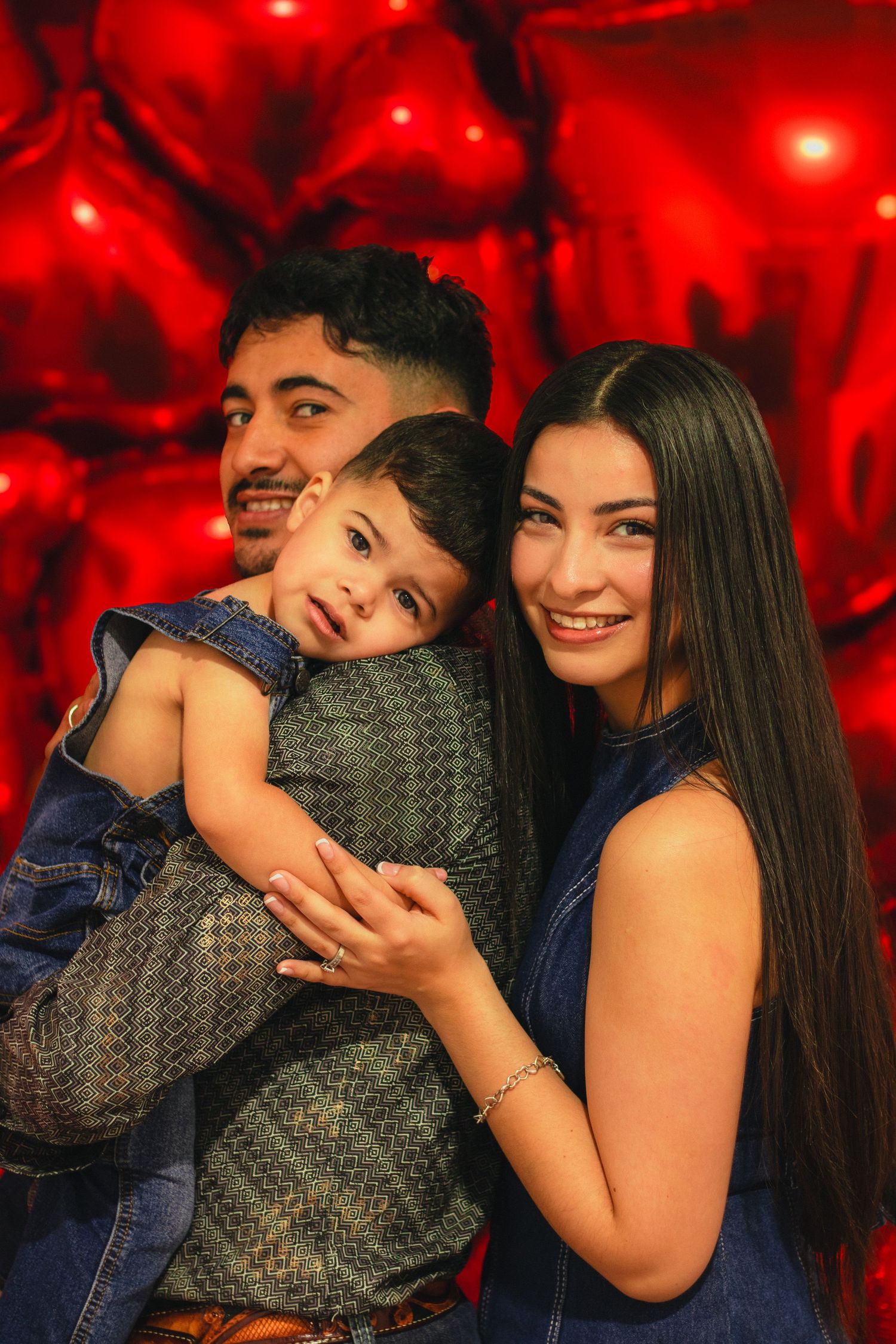 Happy family portrait with toddler between parents against red heart balloons.