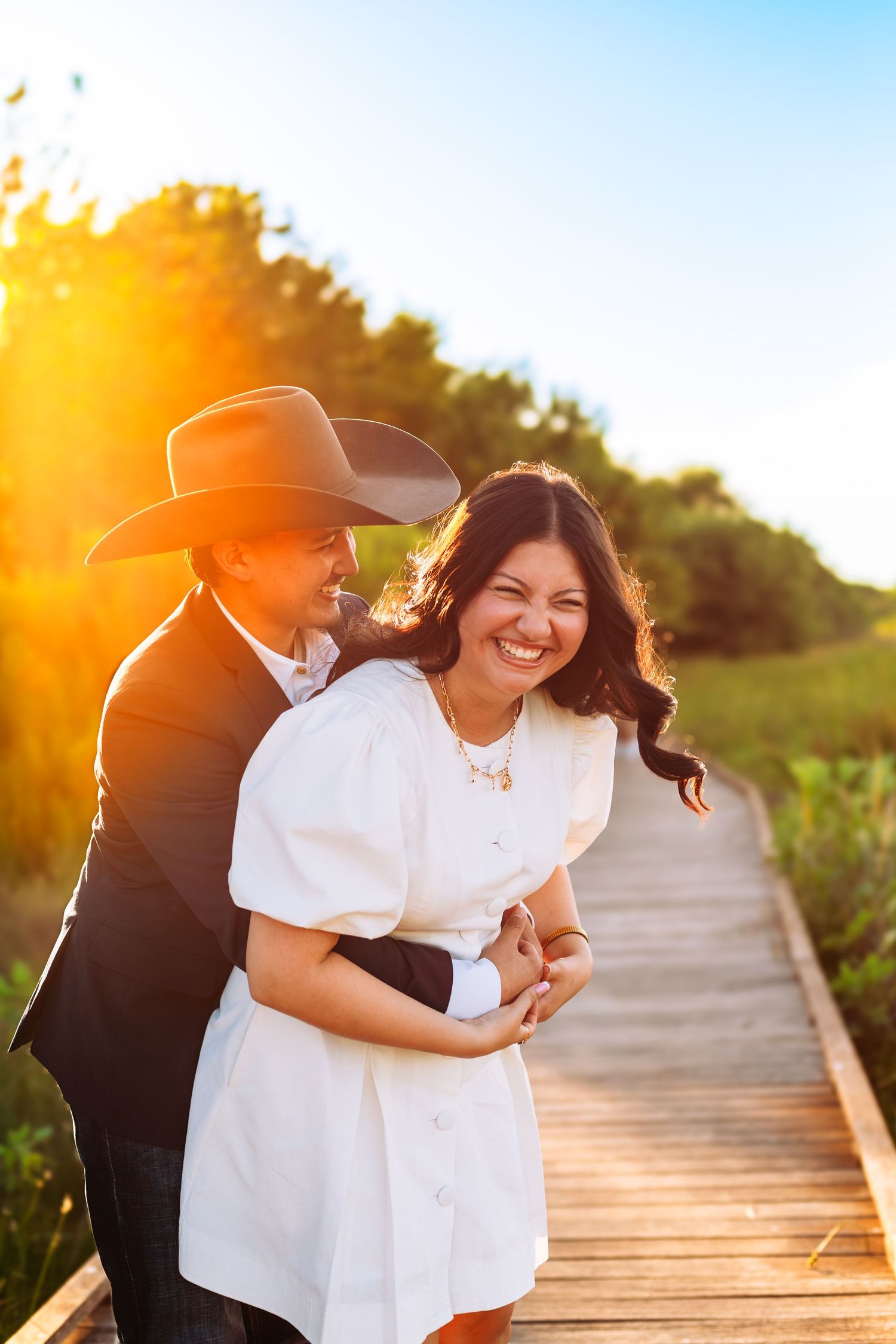 Couple embracing on wooden boardwalk at golden sunset, woman in white dress laughing, man in cowboy hat hugging her.