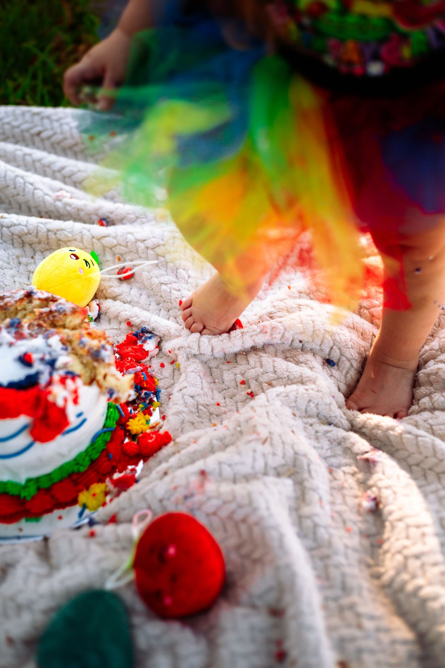 Toddler smashing colorful rainbow birthday cake on white blanket outdoors during first birthday cake smash celebration.