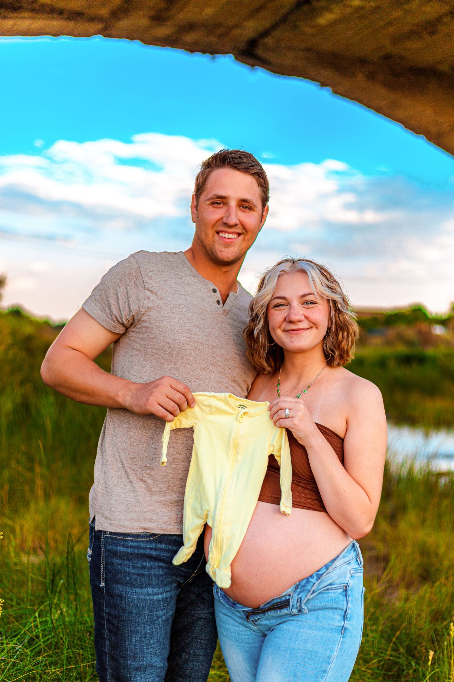 Smiling couple holding yellow baby outfit outdoors, pregnant woman showing baby bump, standing under bridge with blue sky.