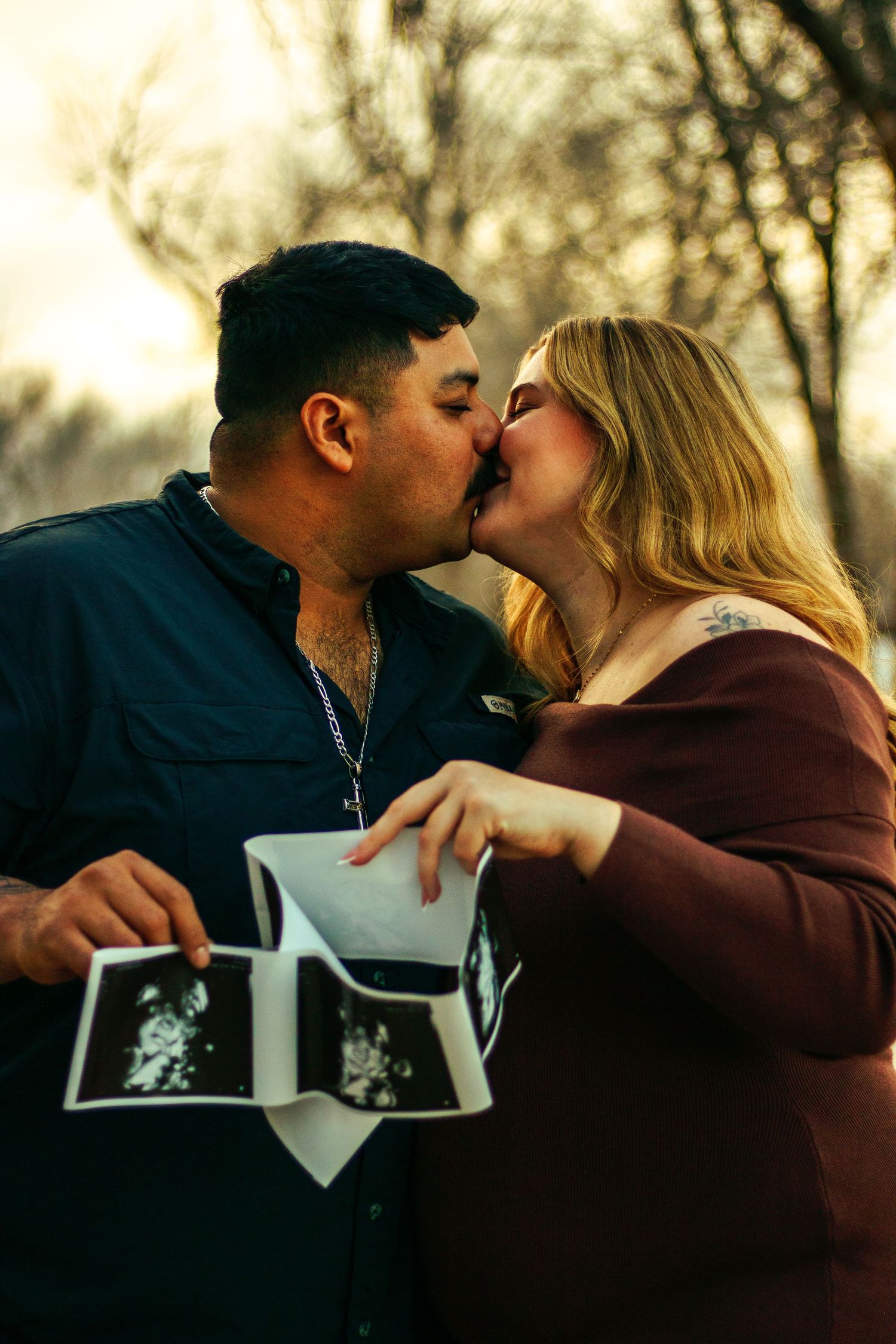Couple kissing outdoors while holding ultrasound photos, announcing pregnancy in a romantic sunset setting.