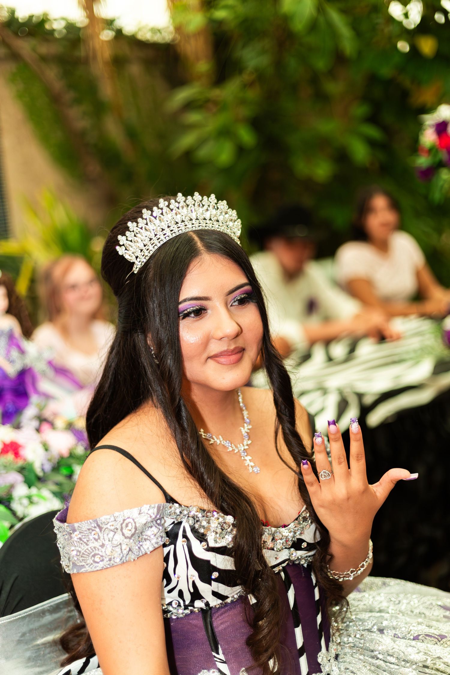 Young woman in a sparkling tiara and elegant off-shoulder gown waves at a quinceañera celebration outdoors.