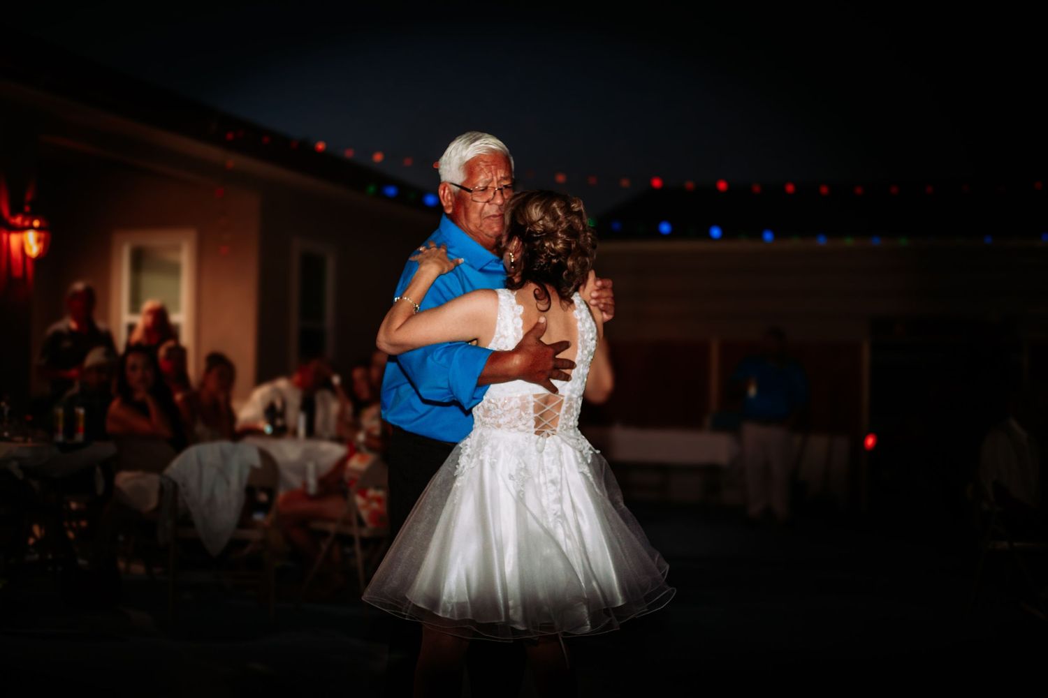 Bride and older man share an emotional father-daughter dance at an outdoor evening wedding reception with colorful string lights.