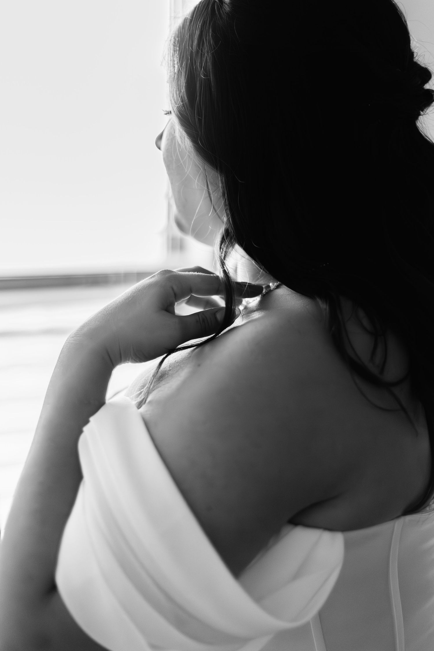 Black and white photo of a bride in a white off-shoulder gown looking out a window on her wedding day.