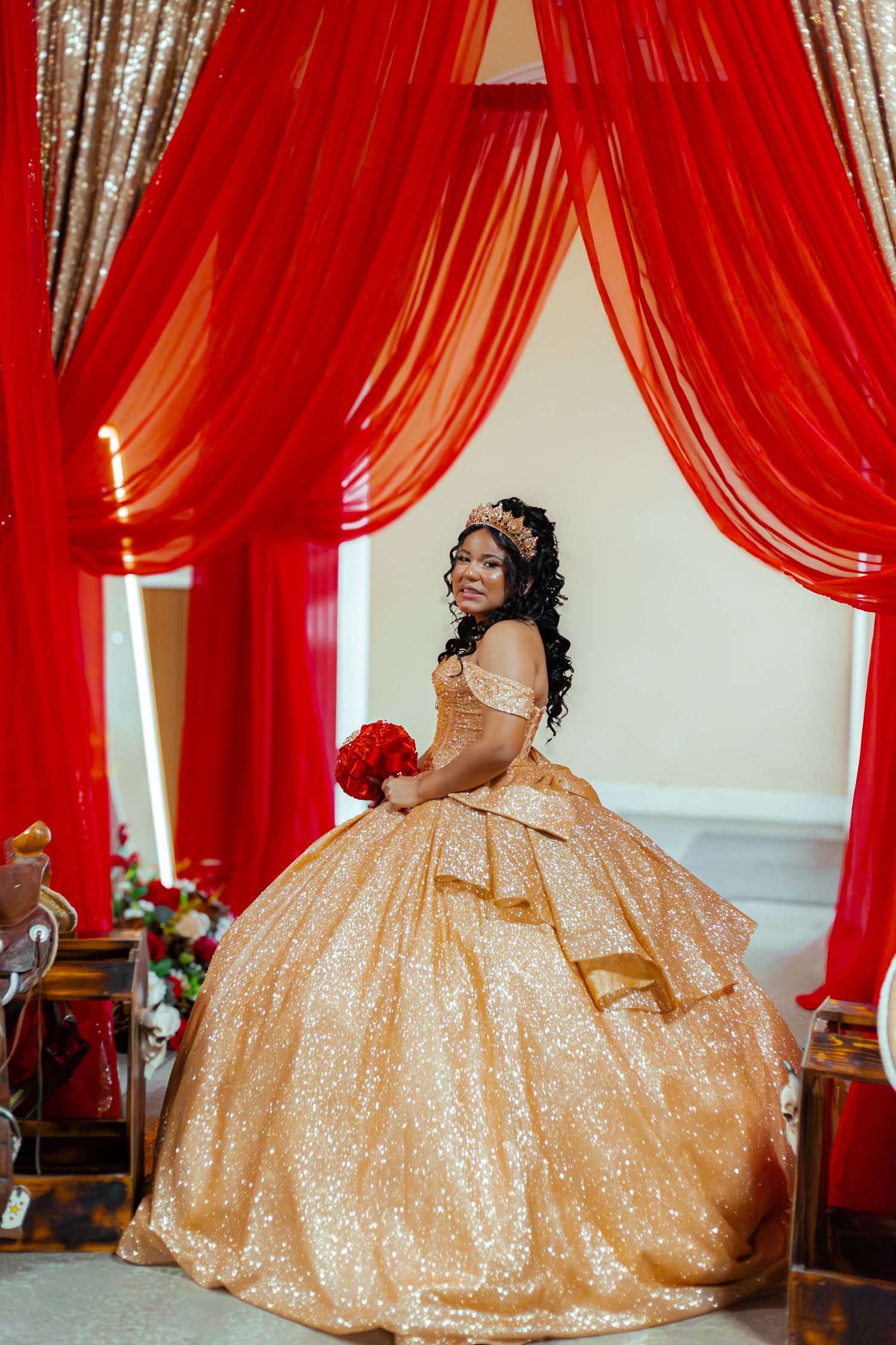 Young woman in gold sequin ball gown with tiara holds red bouquet at quinceañera celebration.