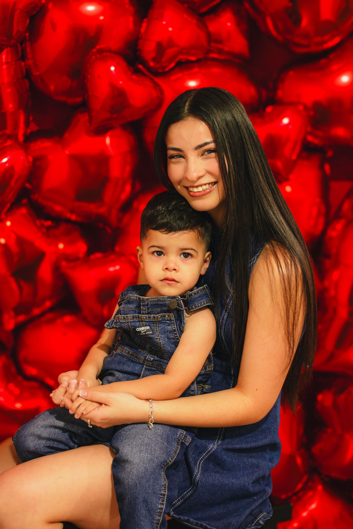 Smiling mother holding toddler in denim outfit against wall of red heart-shaped balloons.