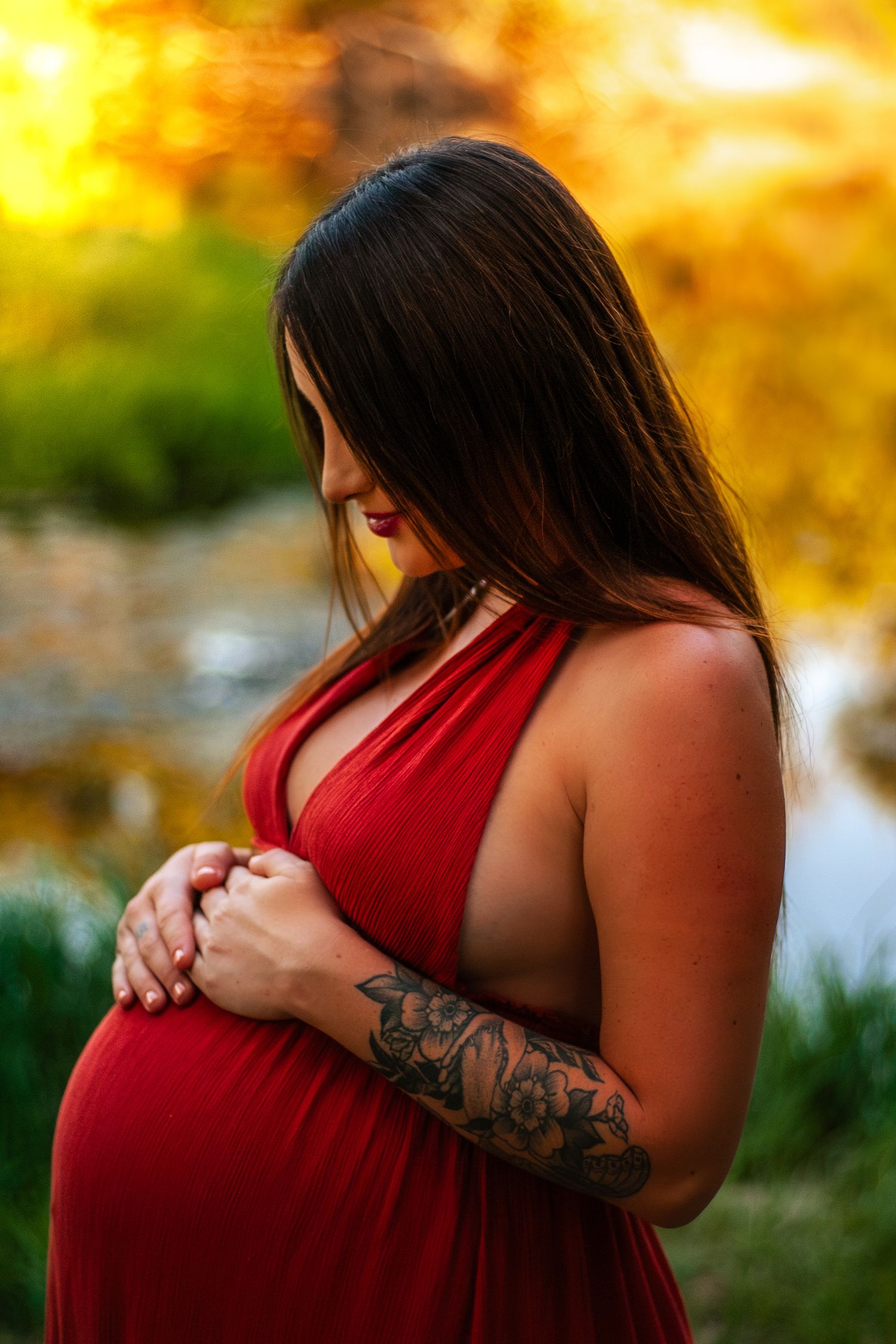 Pregnant woman in red halter dress with floral tattoo gently holds her baby bump near a golden pond.