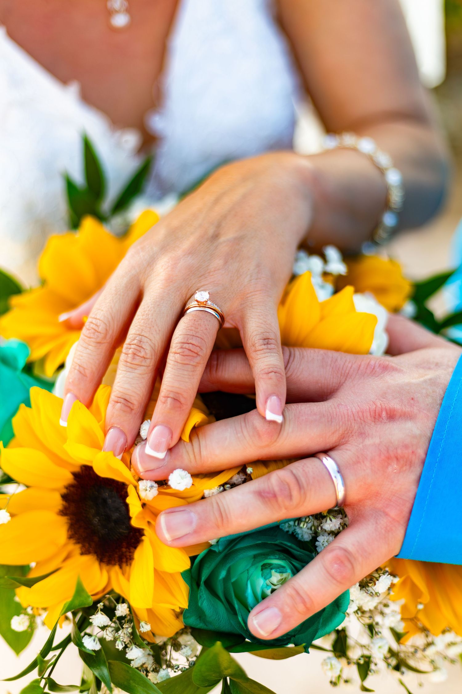 Newlywed couple displaying wedding rings on hands resting over a vibrant sunflower bouquet with teal roses.