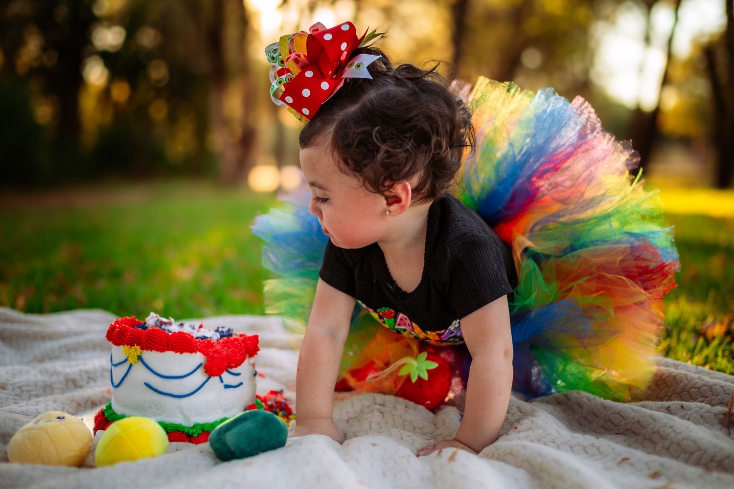 A toddler in a rainbow tutu and red bow headband plays on a white blanket in a sunny park setting.