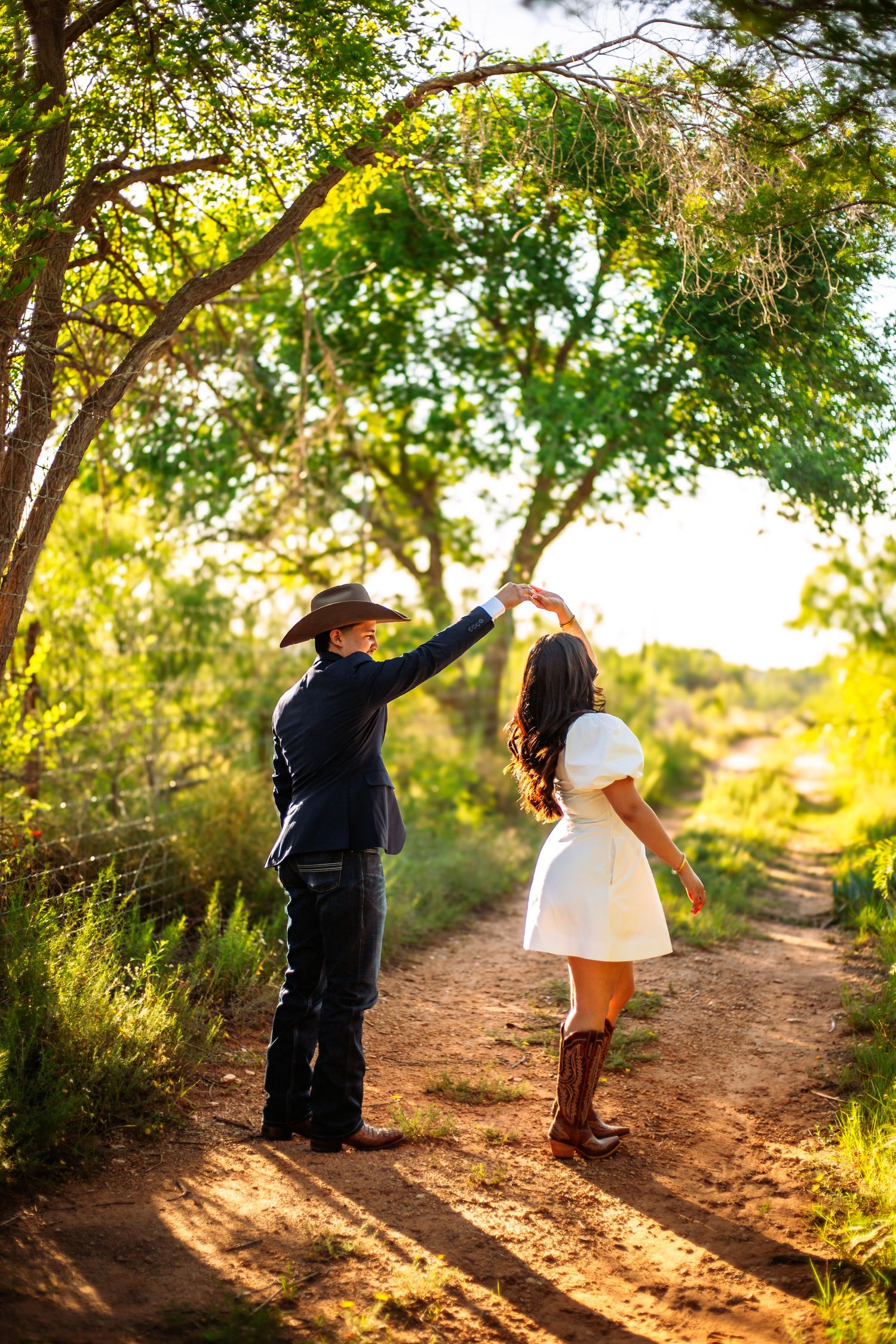 Couple dancing on a sunlit dirt path, man in cowboy hat and woman in white dress with boots, surrounded by lush green trees.
