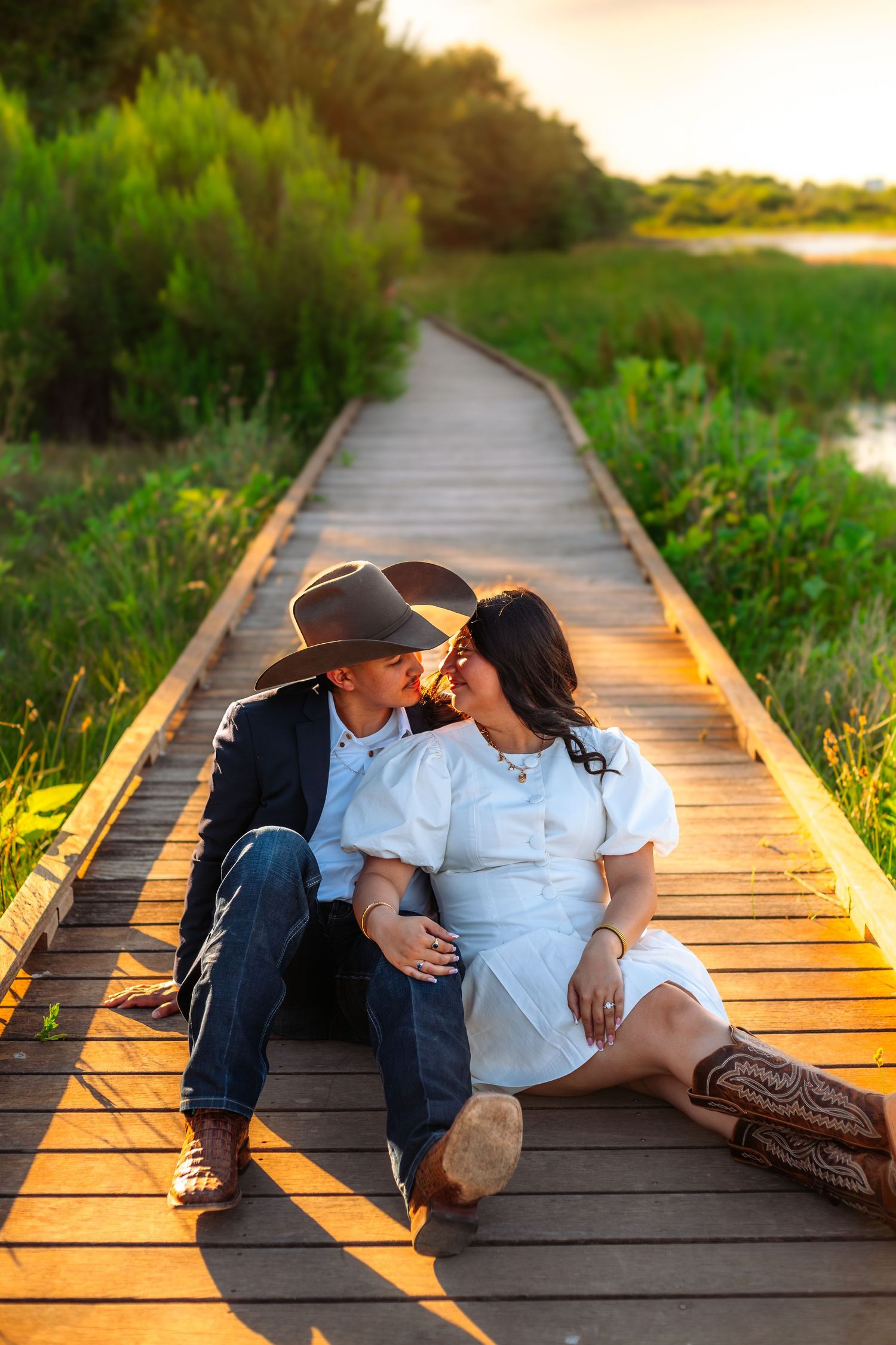 Couple kissing on a wooden boardwalk at sunset, surrounded by lush greenery, man in cowboy hat, woman in white dress.
