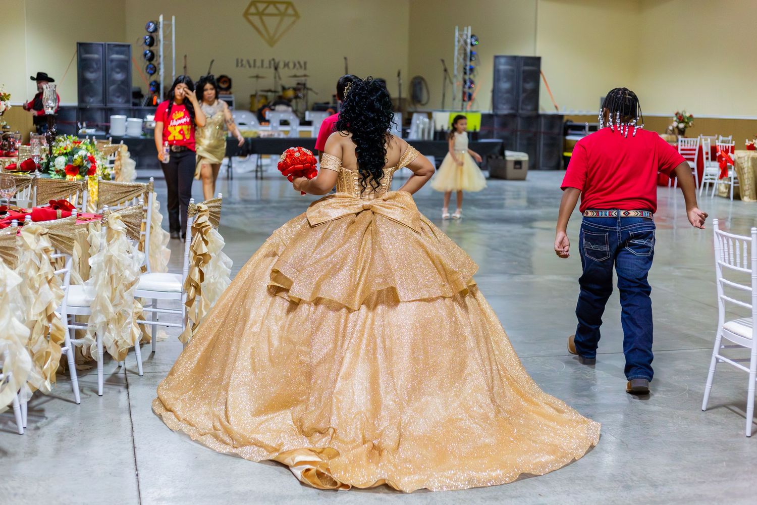Young woman in elegant gold quinceañera ballgown walks through decorated event hall during celebration.