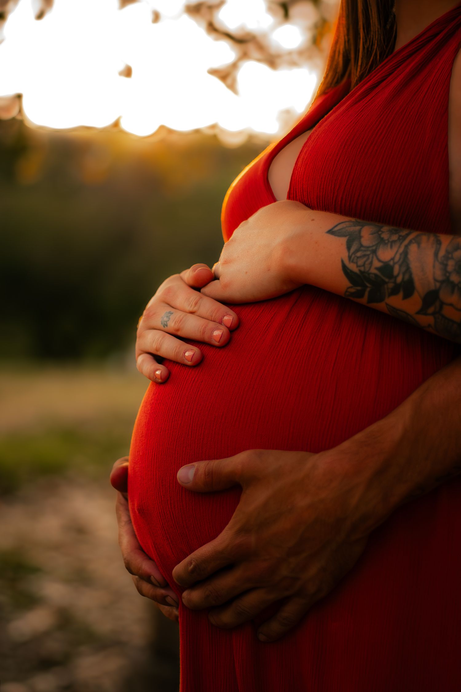 Close-up of tattooed pregnant couple tenderly holding baby bump in red dress at sunset.