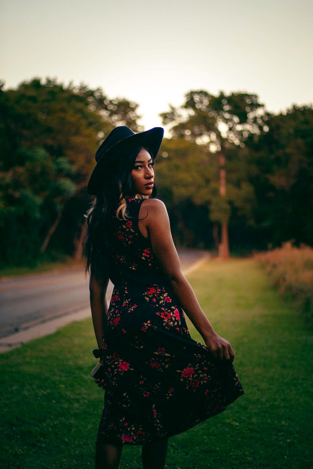 Young woman in floral dress and black hat standing on grass at sunset near a road.