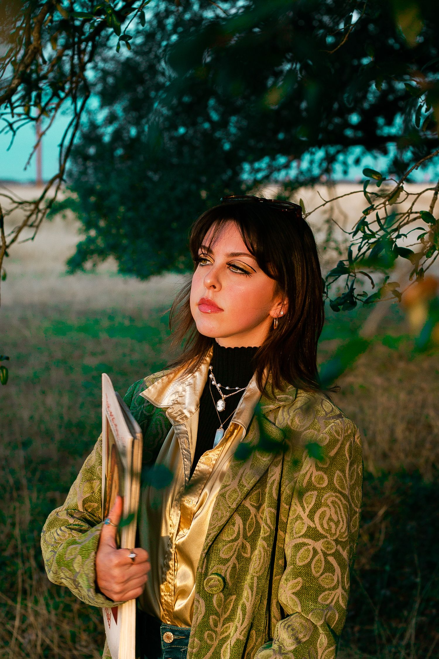 Dark-haired woman in gold jacket holding a shovel under a tree at sunset.