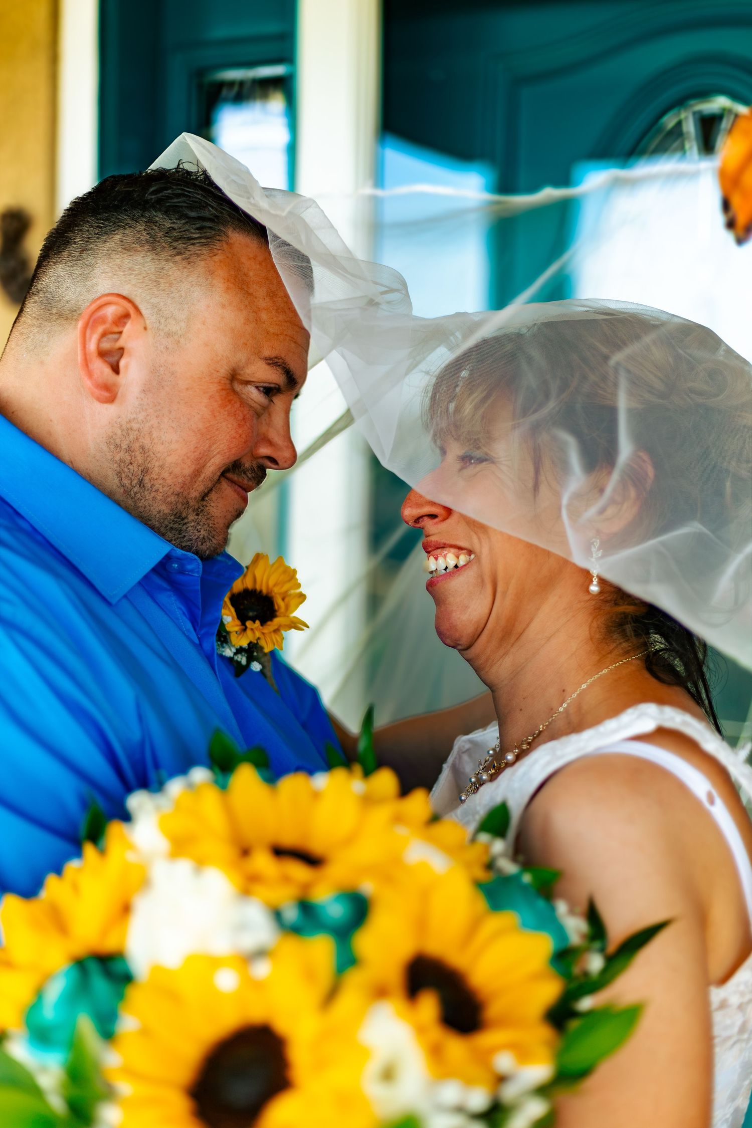 Bride and groom share a romantic moment under a veil, holding a sunflower bouquet on their wedding day.