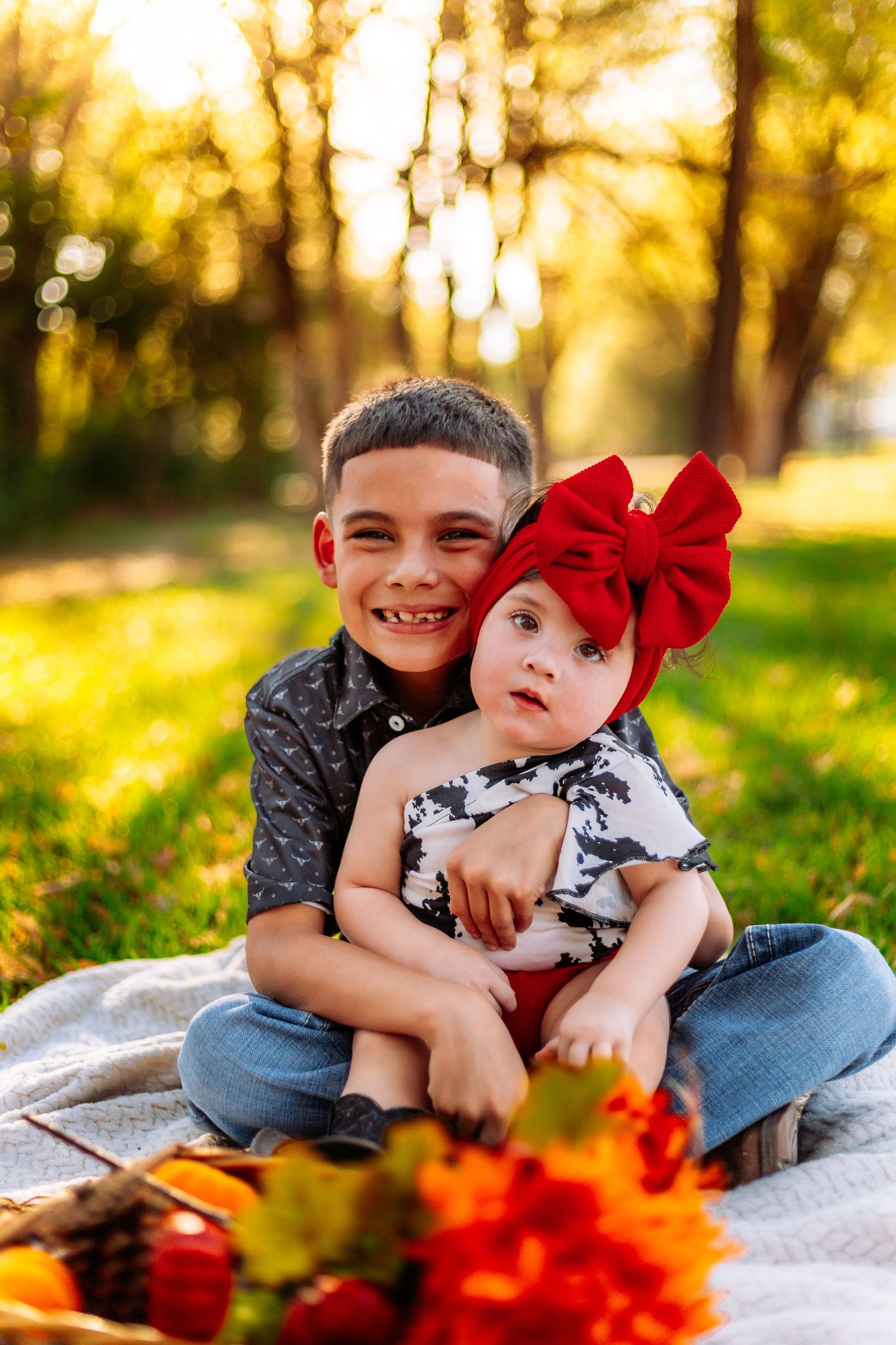 Boy hugging baby girl with red bow headband on blanket in autumn park during golden hour sunset.