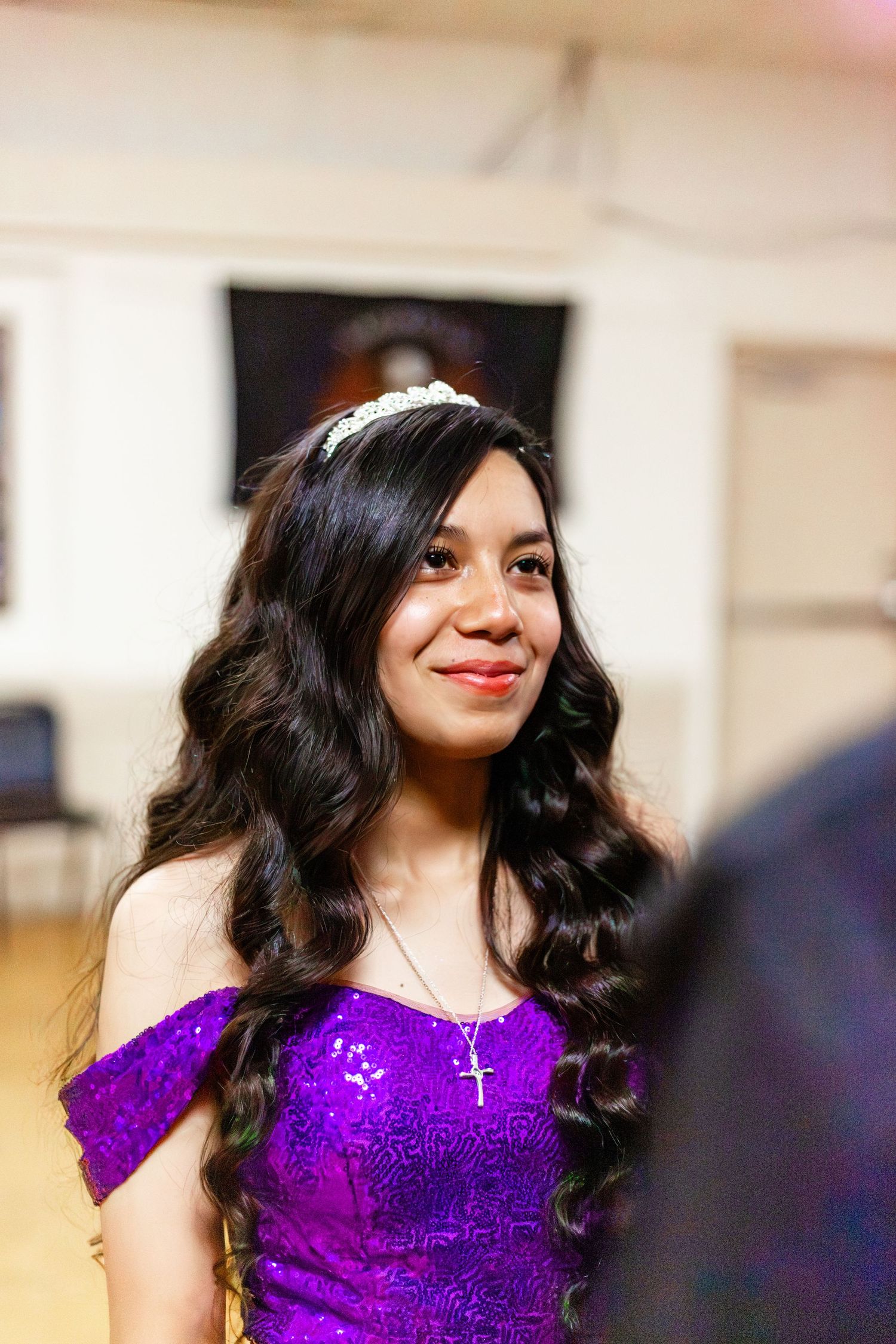 Young woman in purple sequin off-shoulder gown wearing tiara and cross necklace smiling at quinceañera celebration.