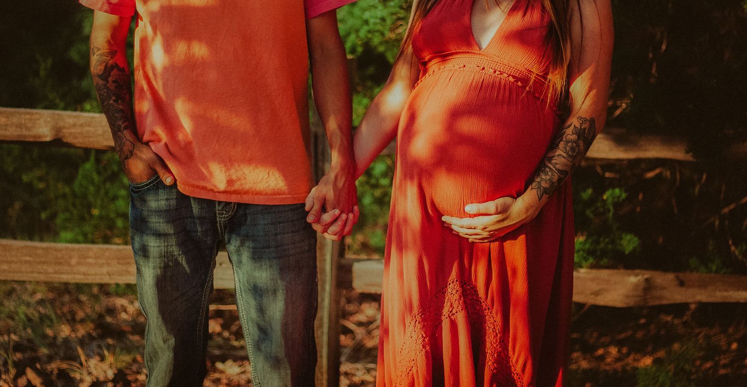 Couple holding hands outdoors, woman in orange maternity dress showing baby bump near wooden fence.