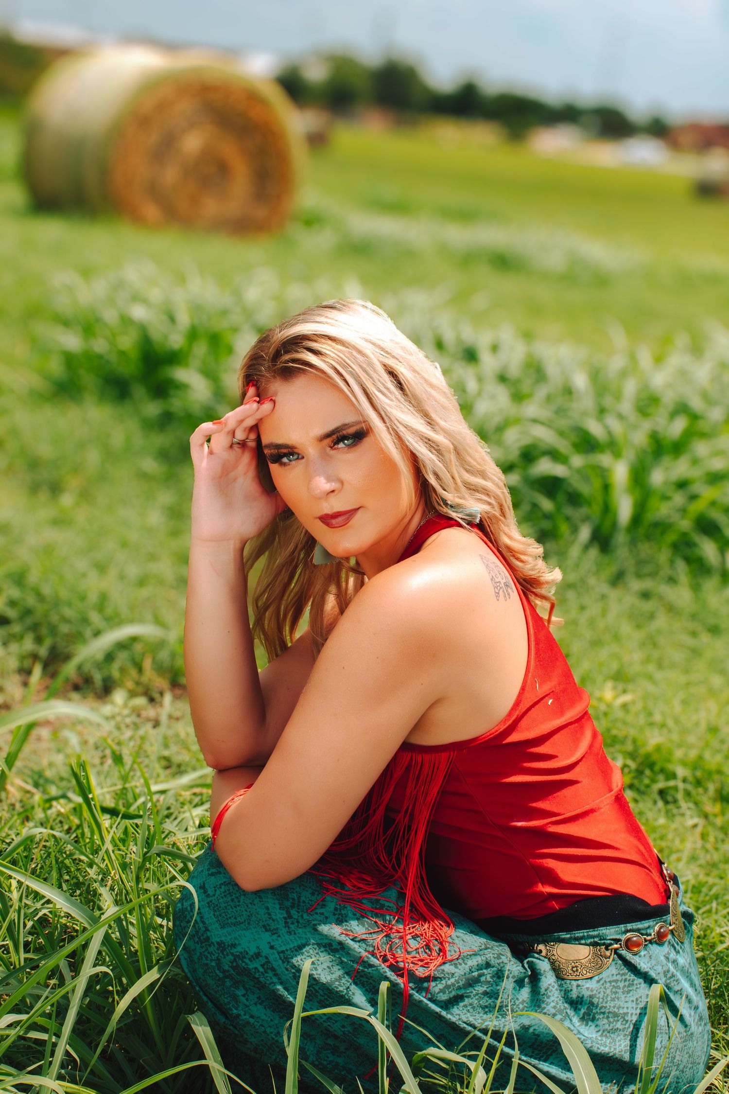 Blonde woman in red top and teal skirt sits in green field with hay bale backdrop.