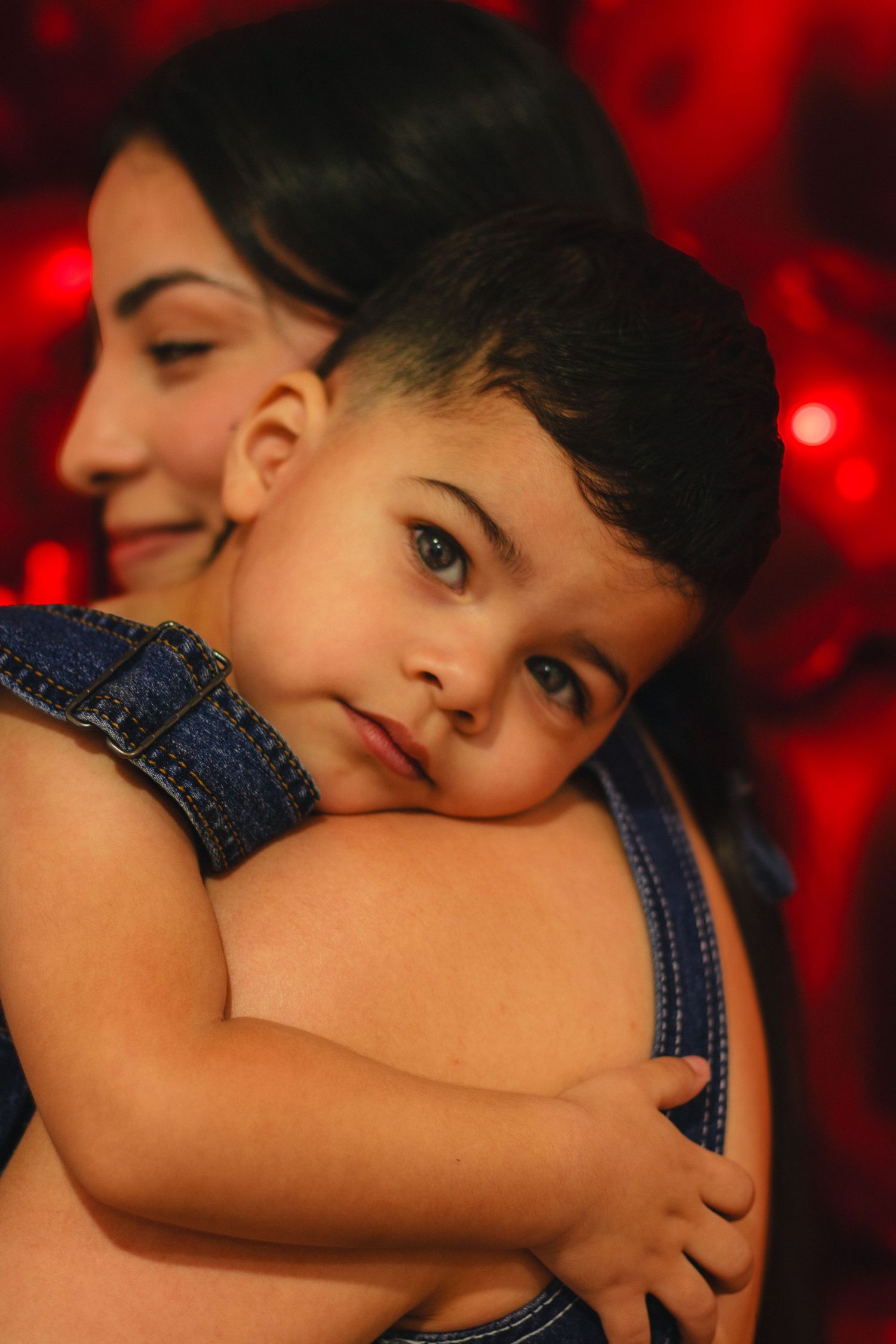Young child in denim overalls hugging mother against red balloon backdrop.