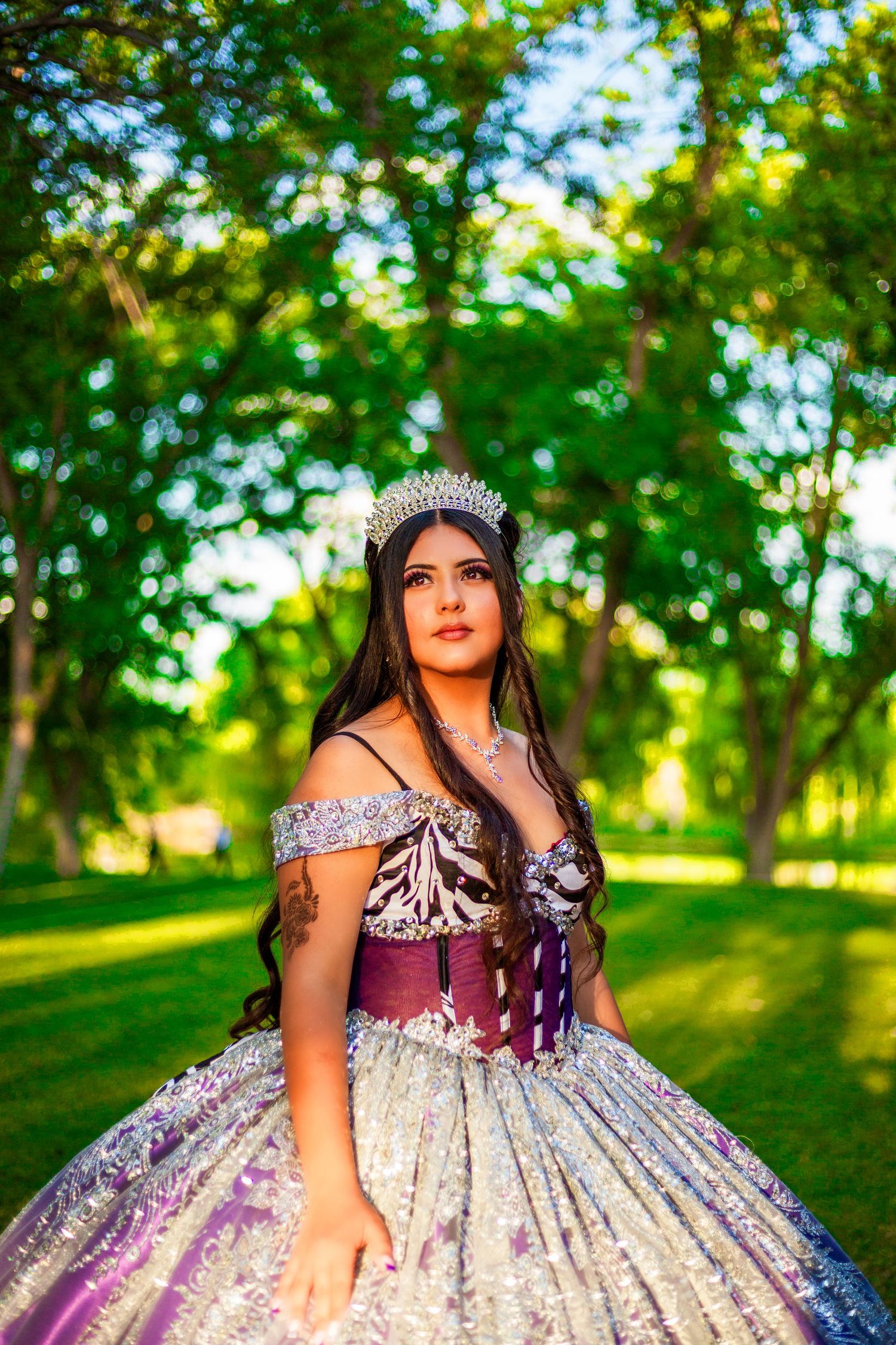 Young woman in a stunning silver and purple quinceañera ball gown with a tiara, posing outdoors among green trees.