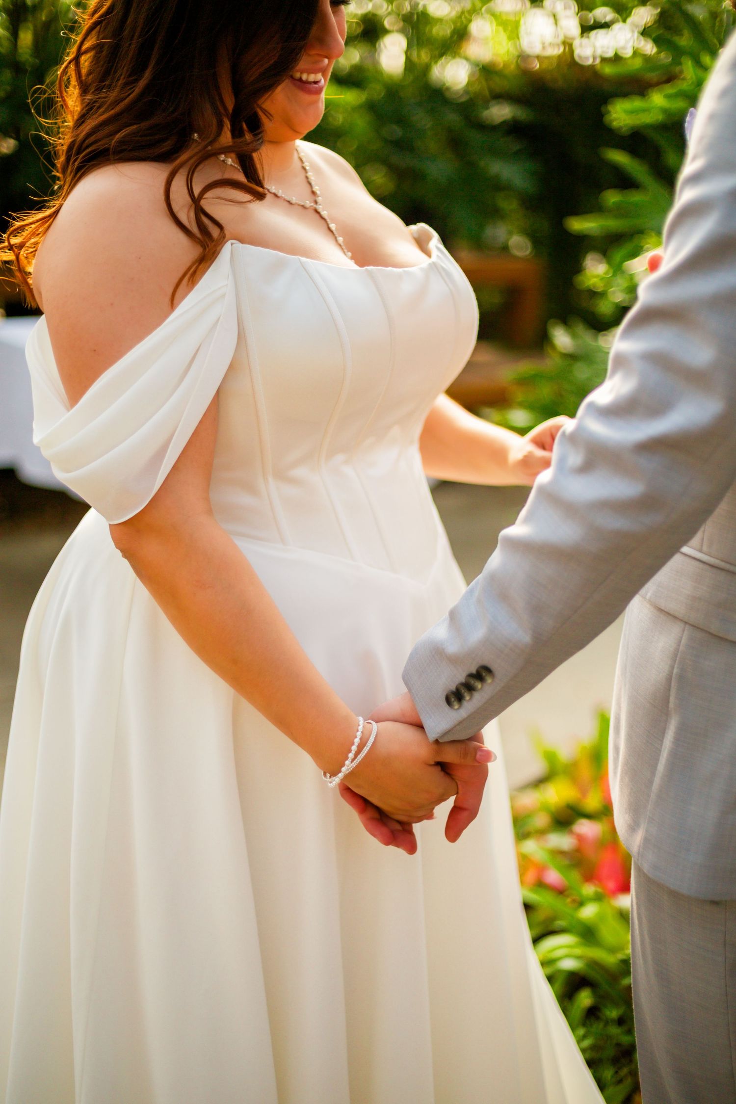 Bride in off-shoulder white gown holds hands with groom in gray suit outdoors at wedding ceremony.