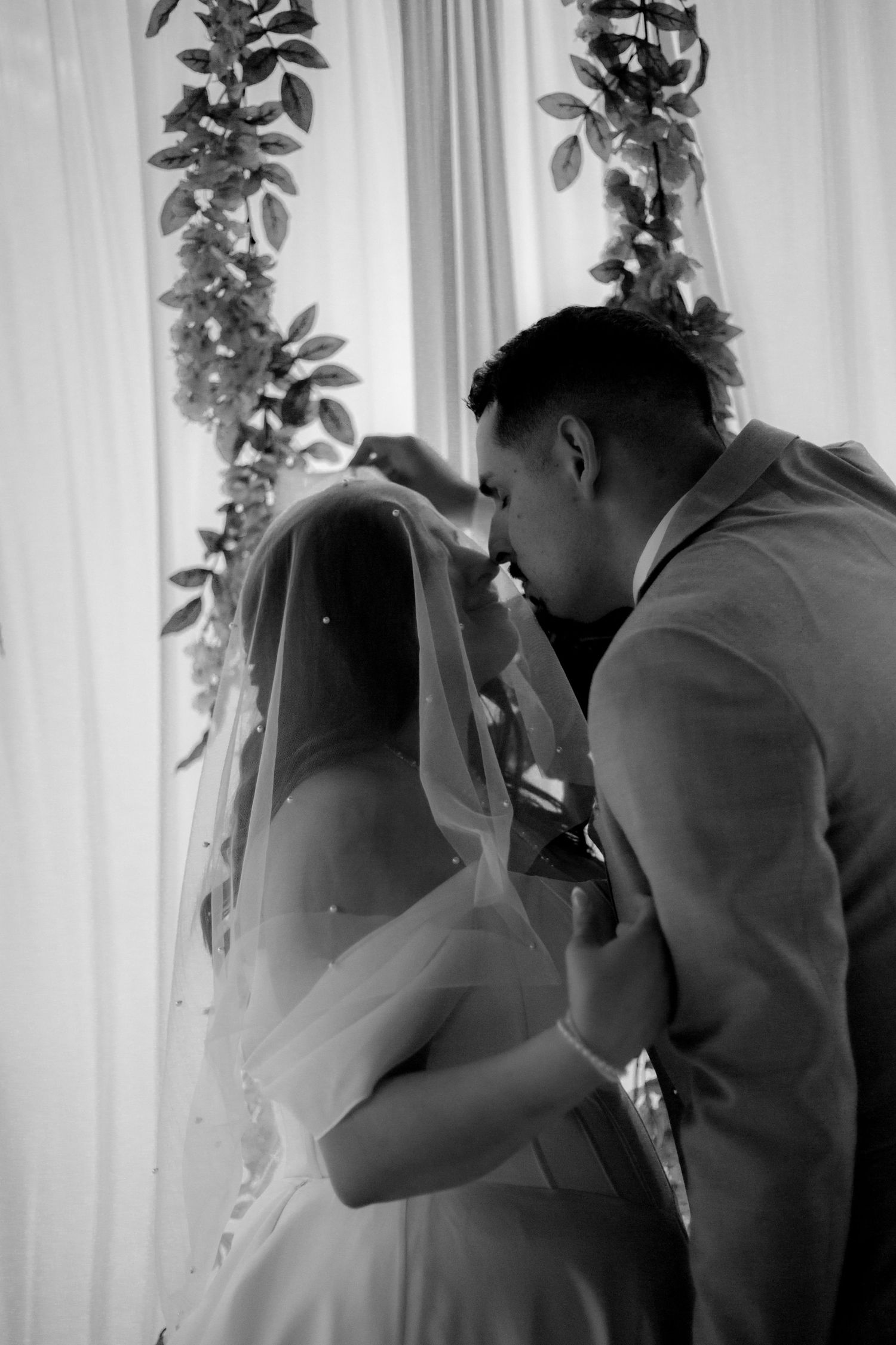 Black and white photo of a groom kissing his bride's forehead during a wedding ceremony with floral decor.