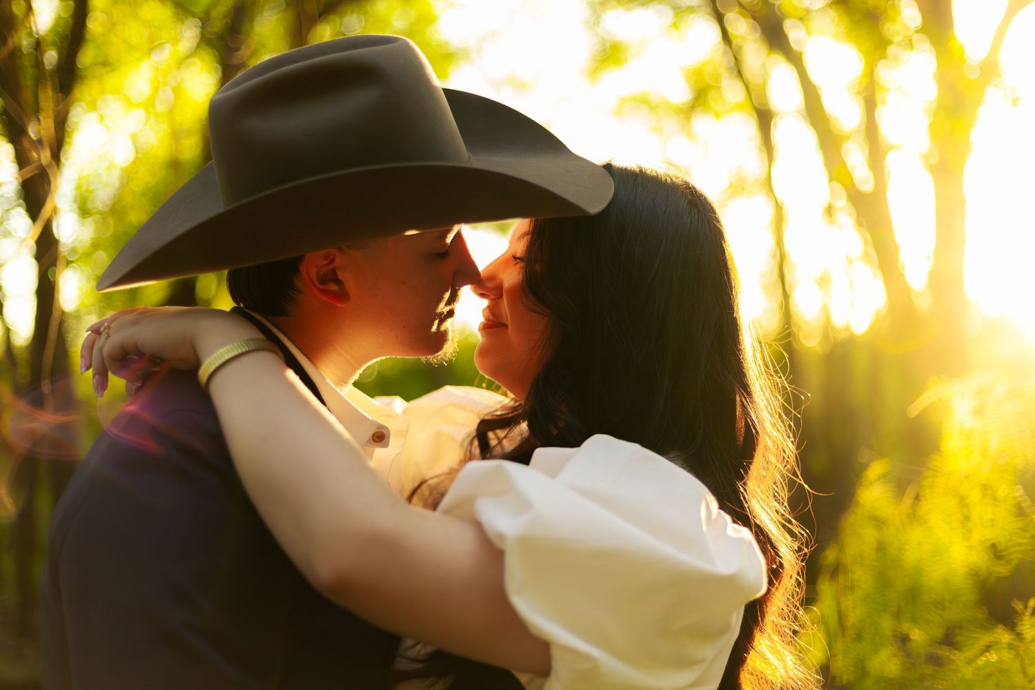 Couple sharing a romantic moment at sunset, man wearing a cowboy hat embracing woman in golden forest light.
