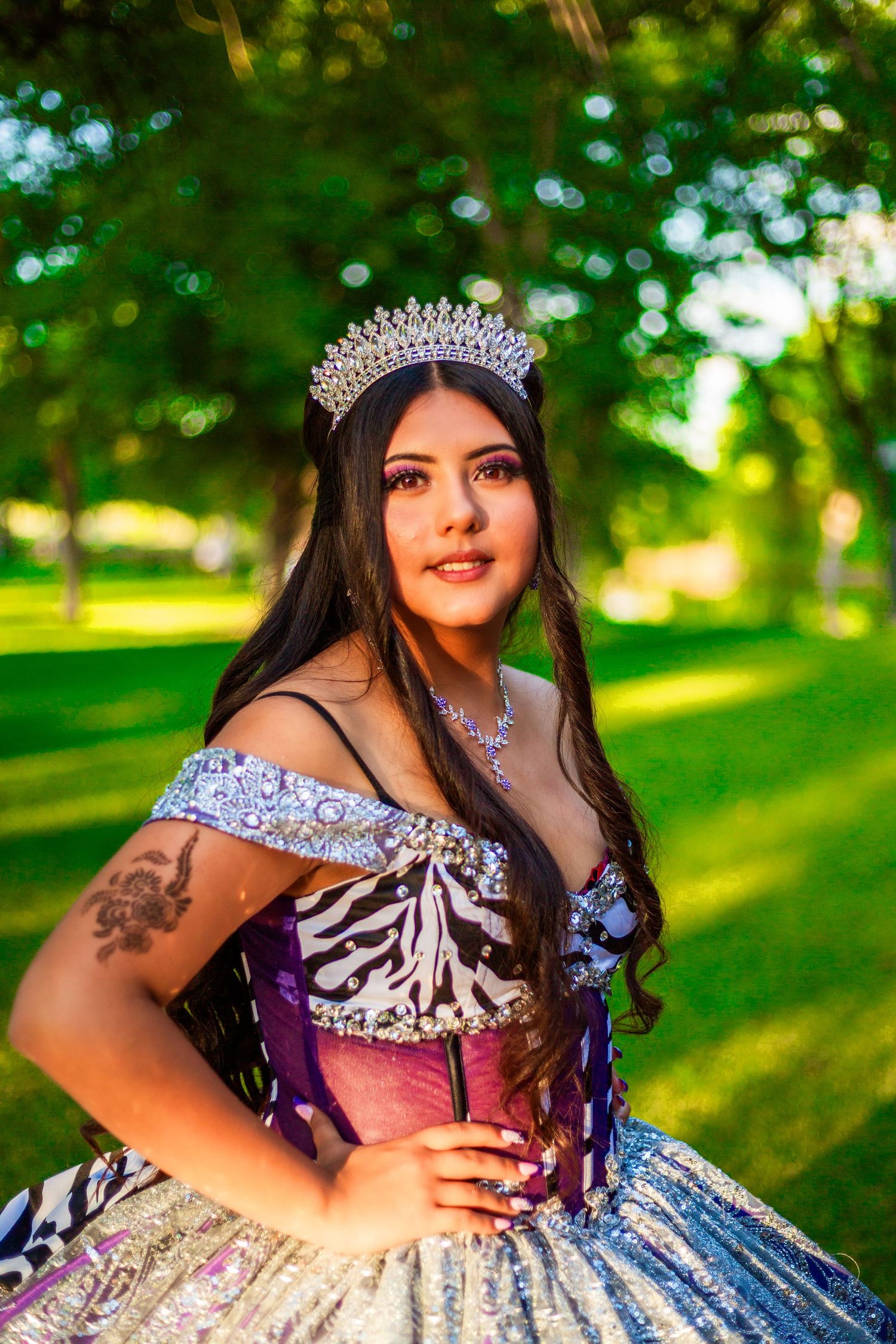 Young woman in a sparkling quinceañera gown and tiara poses gracefully in a lush green park.