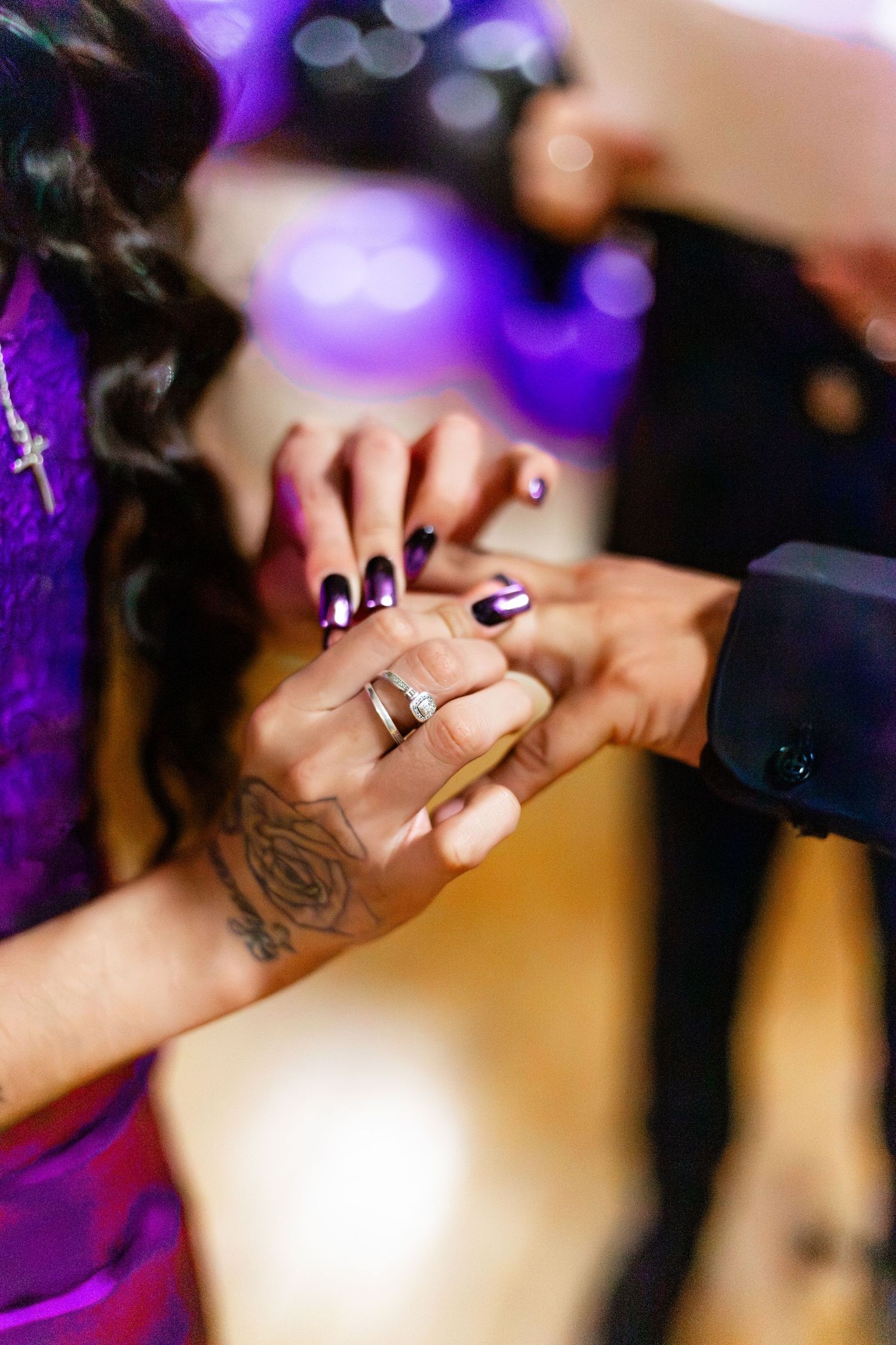 Close-up of woman's hand with diamond engagement ring and rose tattoo held by man in black suit.