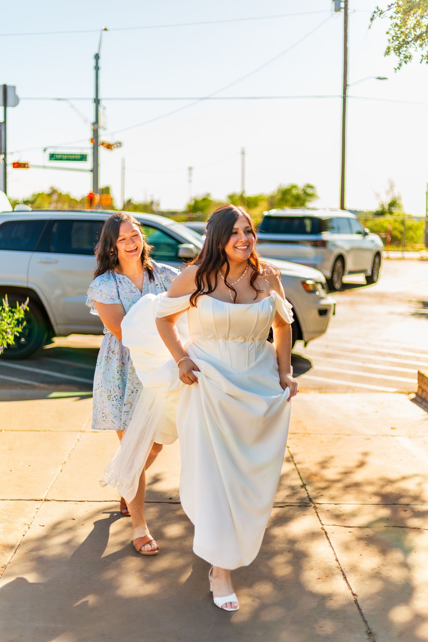 Two women in elegant dresses walk together on a sunny sidewalk, smiling joyfully near a parking lot.