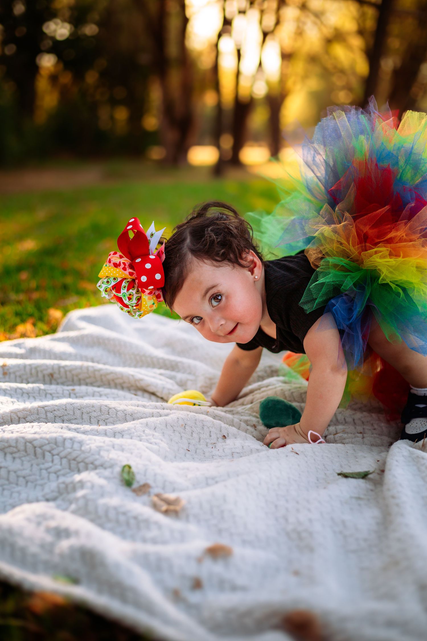 Baby girl in colorful rainbow tutu and red bow crawls on white blanket in sunny autumn park.