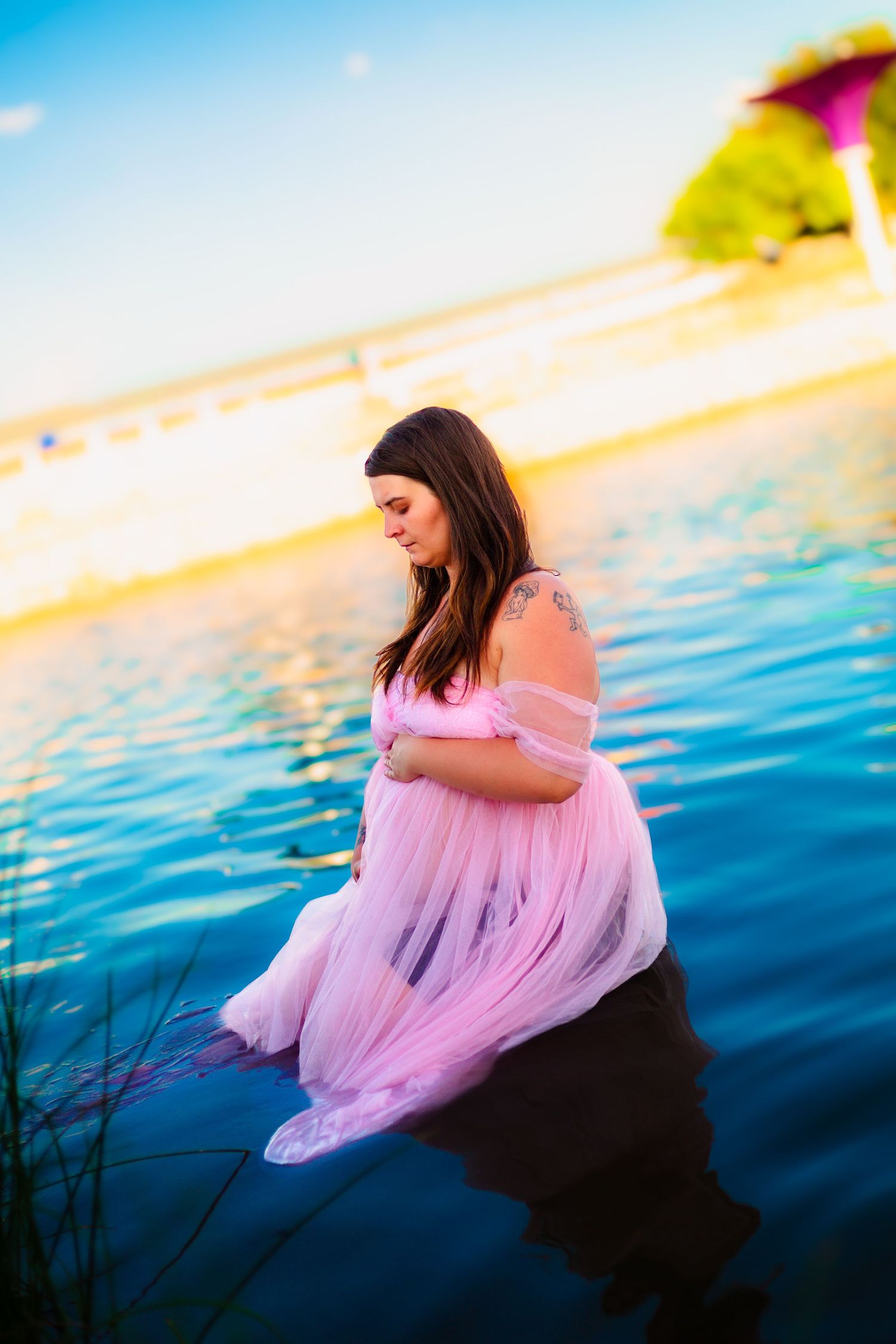 Pregnant woman in pink tulle dress standing in calm blue water during golden hour at a lakeside beach.