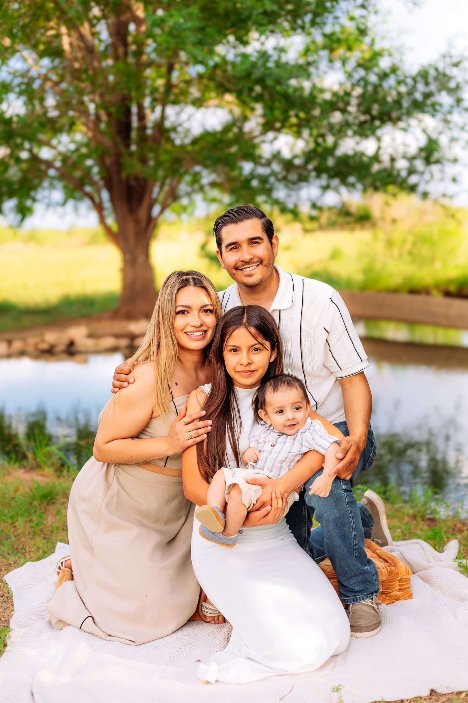 Happy family of four posing outdoors near a pond, with parents and two children sitting on a blanket in a park.