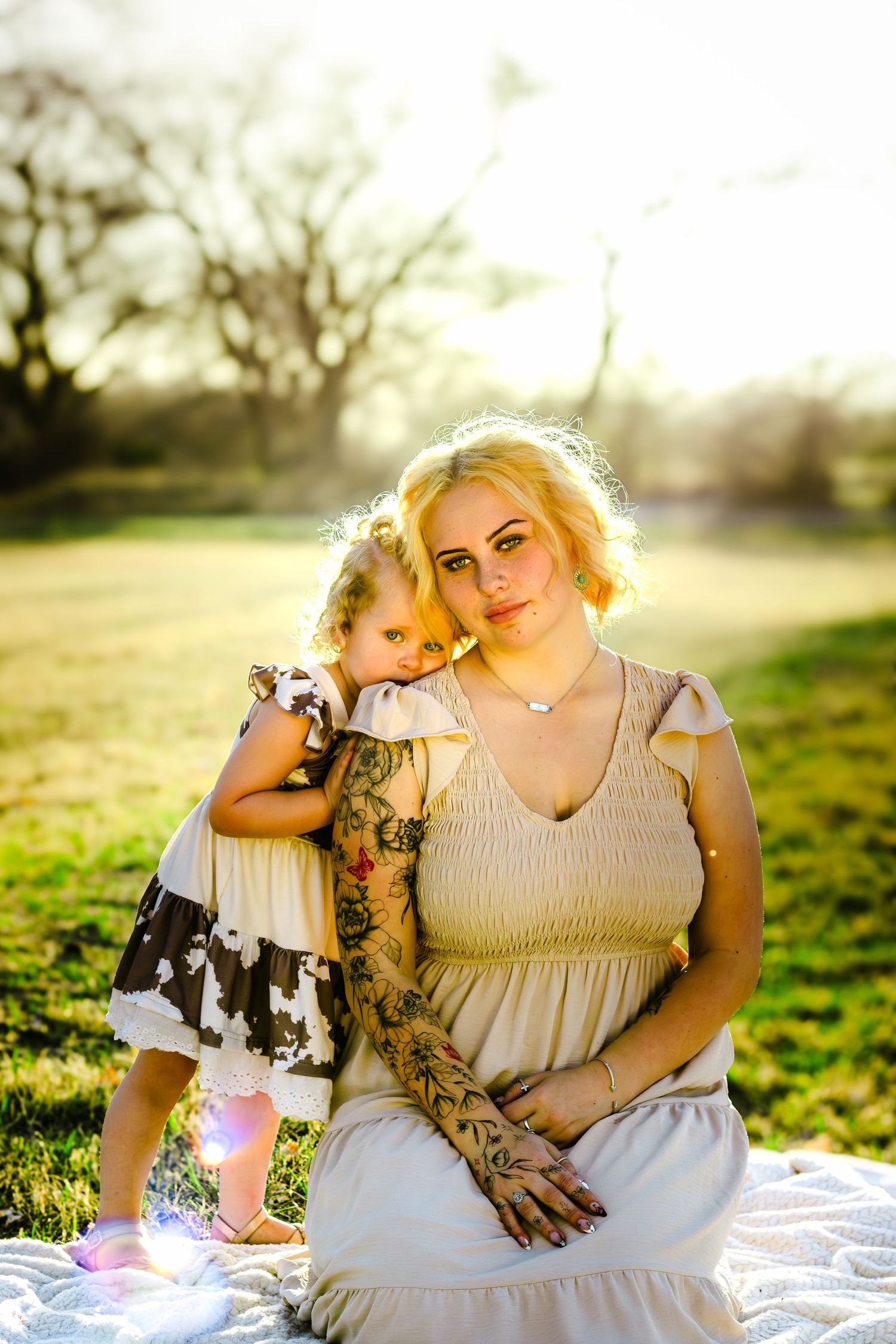 Tattooed blonde mother sitting with young daughter in sunny outdoor park.